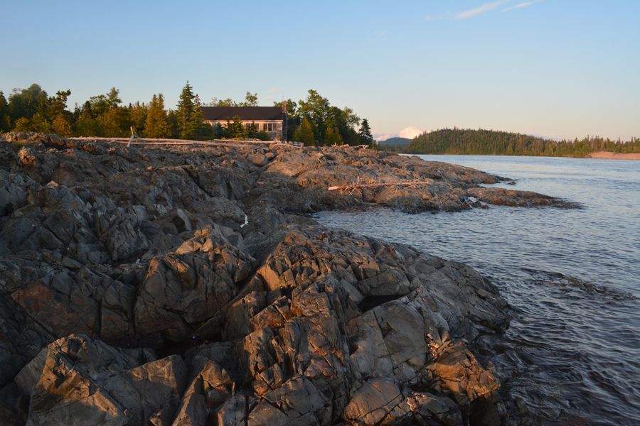 Rotsachtige oever van een meer bij zonsondergang met kalm water, bomen in de verte het hoofdgebouw van Rock Island Lodge aan de horizon.