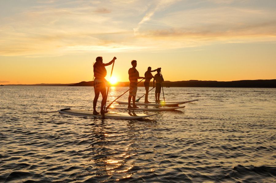 Mensen peddelen op een stand-up paddleboard bij zonsondergang op kalm water, met de oranje lucht als silhouet.