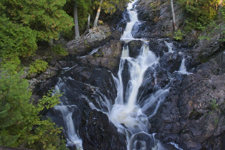 Een waterval die over donkere rotsen naar beneden stort, omgeven door groene bomen en mos.