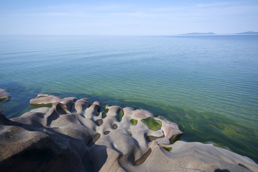 Golven kabbelen tegen de gladde rotsachtige kust onder een kalme blauwe hemel en zee.