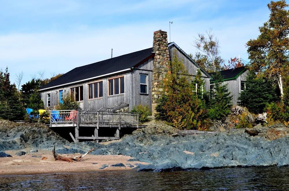 Het hoofdgebouw van Rock Island Lodge aan een rotsachtige kustlijn met bomen en kalm water bij zonsondergang.