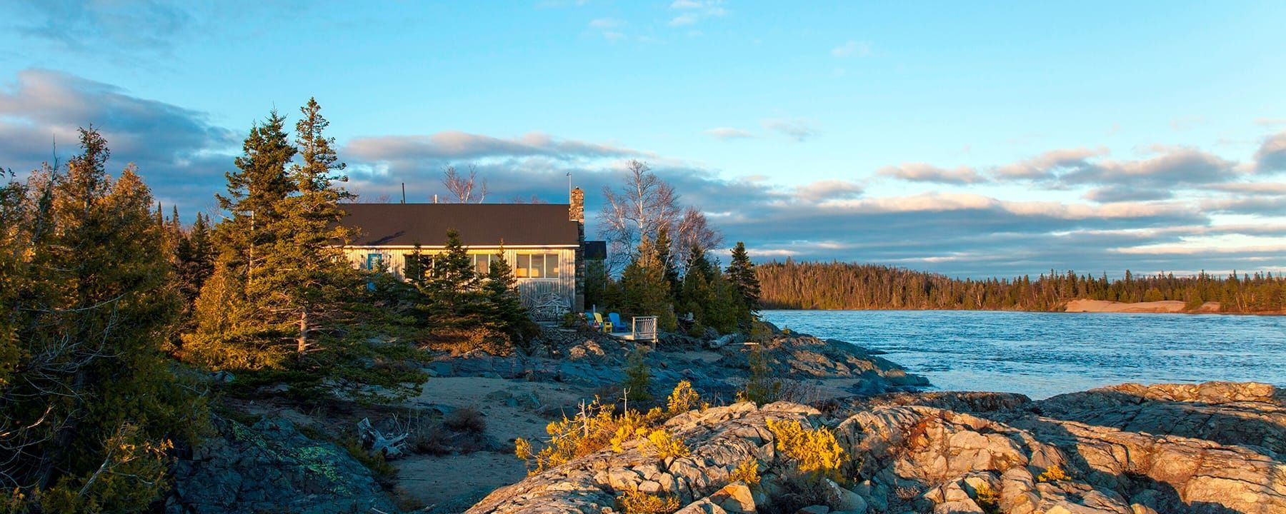 Rock Island Lodge aan de kust bij zonsondergang, gelegen naast een rotsachtige kustlijn, met bomen en kalm water onder een bewolkte hemel.