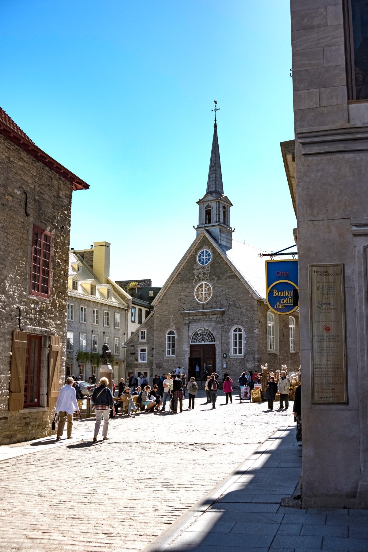 Zonovergoten stenen plein Place Royale in Old Quebec met een kerk en een hoge torenspits; mensen verzamelden zich ervoor op een heldere dag.
