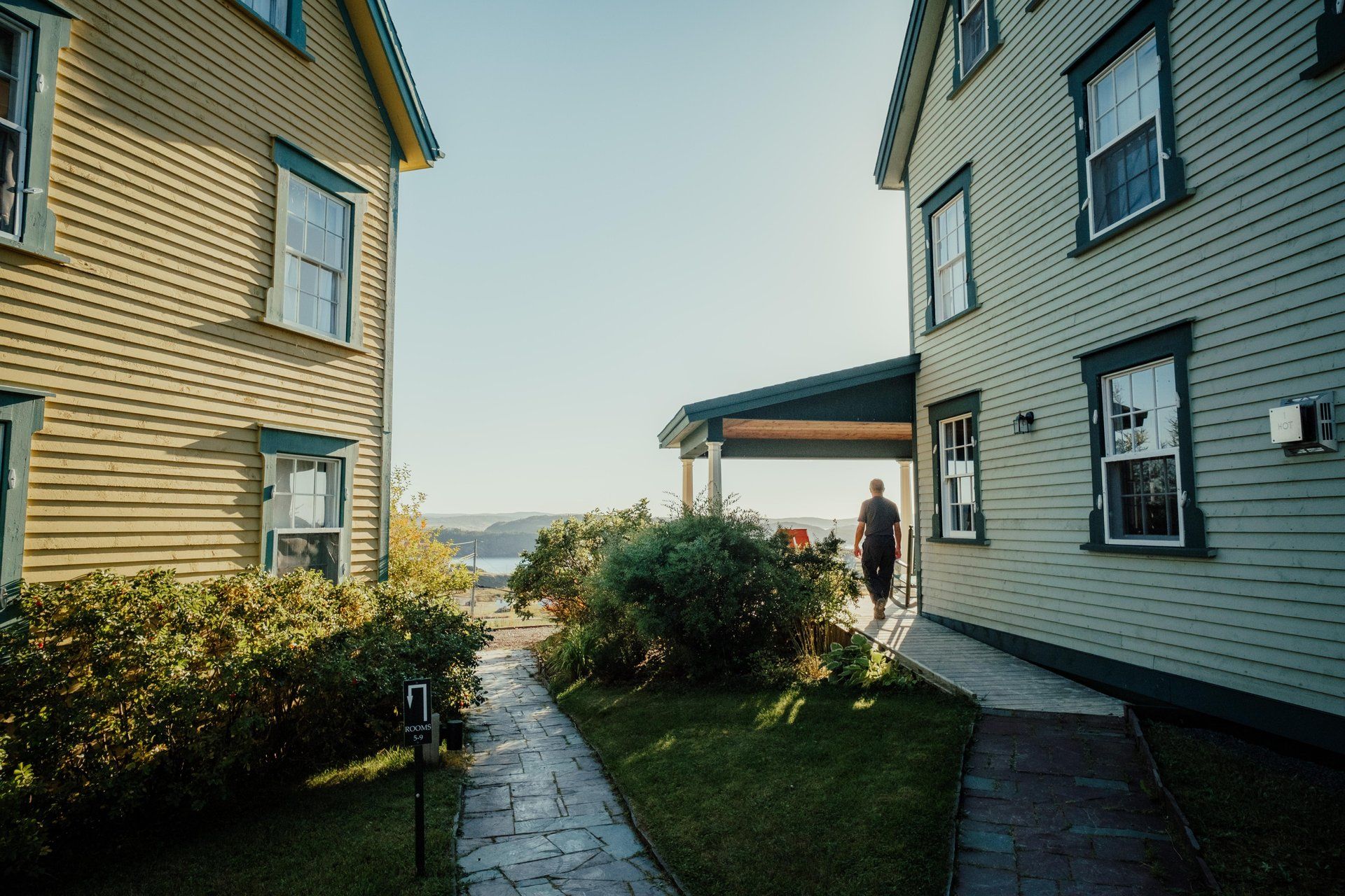 Een smal pad tussen twee huizen van Fisher's Loft Inn in Port Rexton leidt naar een zonnige waterkant, met een persoon die bij de rechter veranda staat.