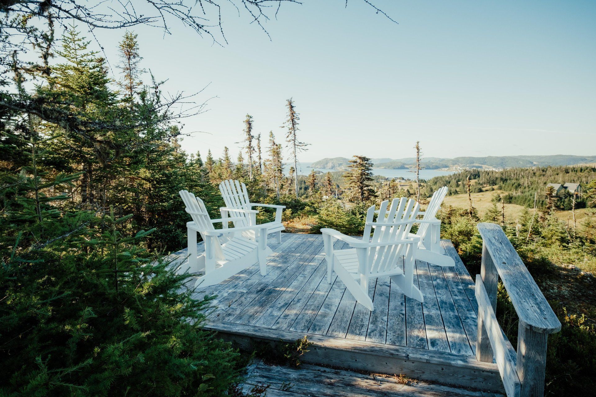 Witte tuinstoelen en een tafel op een houten terras van Fisher's Loft Inn in Port Rexton met uitzicht op een zonnig landschap.