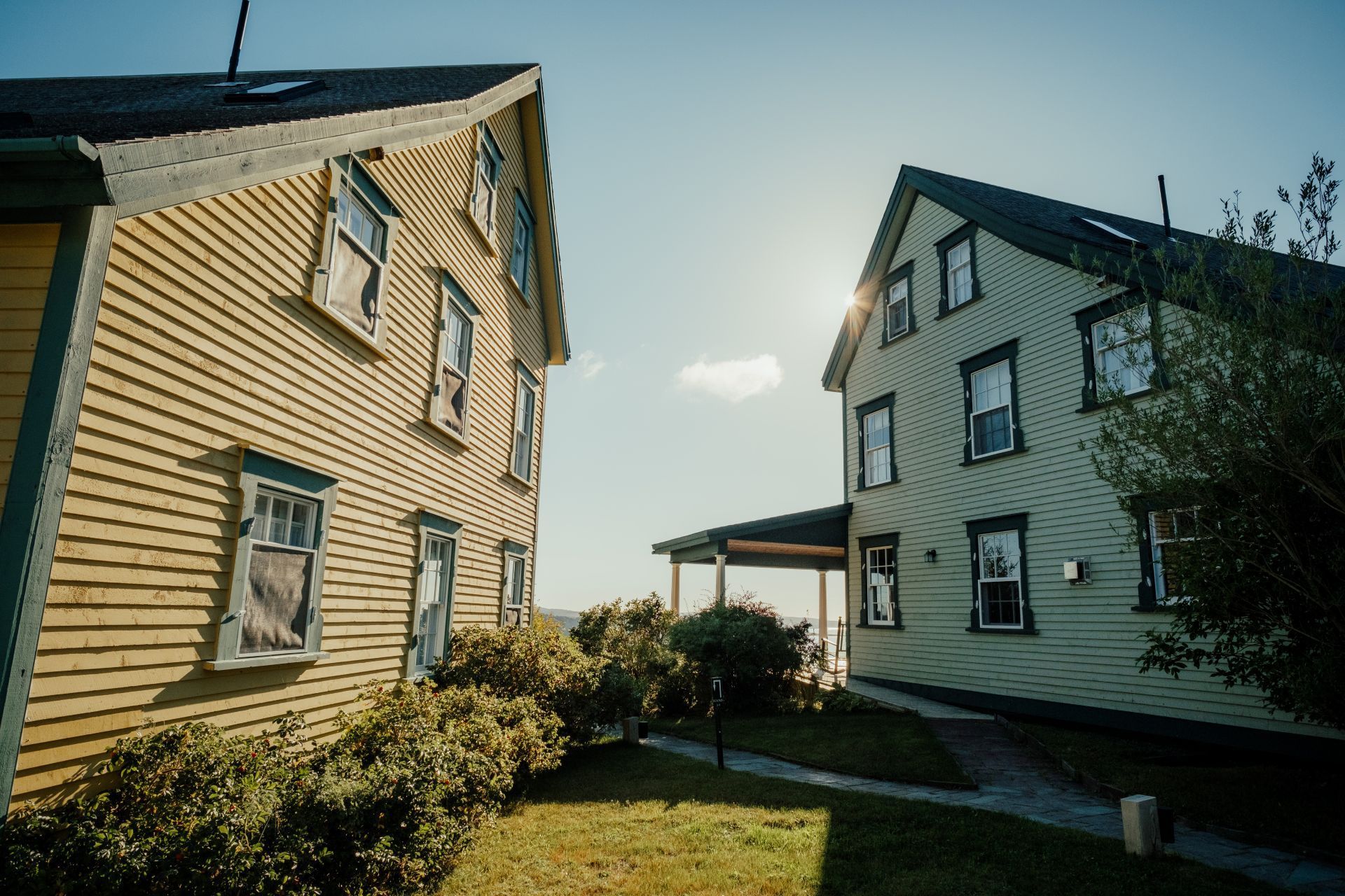 Twee witte huizen van Fisher's Loft Inn in Port Rexton met donkere daken staan tegenover een grasrijke binnenplaats onder een blauwe hemel.