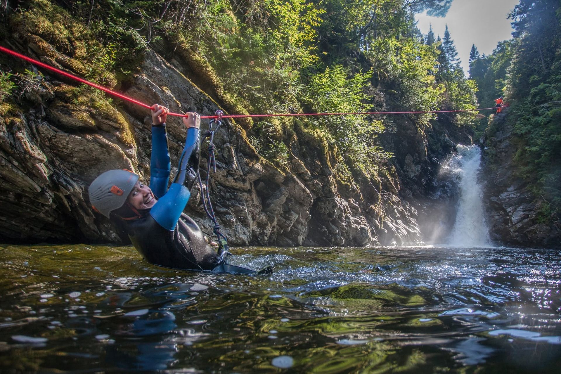 Een persoon in Quebec, Canada, in een blauw pak slingert aan een rood touw boven een rotsachtig watervalbad in een boscanyon.