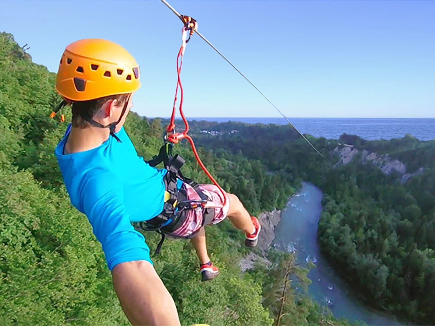 Een klimmer in Quebec, Canada,met helm glijdt via een zipline boven een beboste riviervallei richting de oceaan.