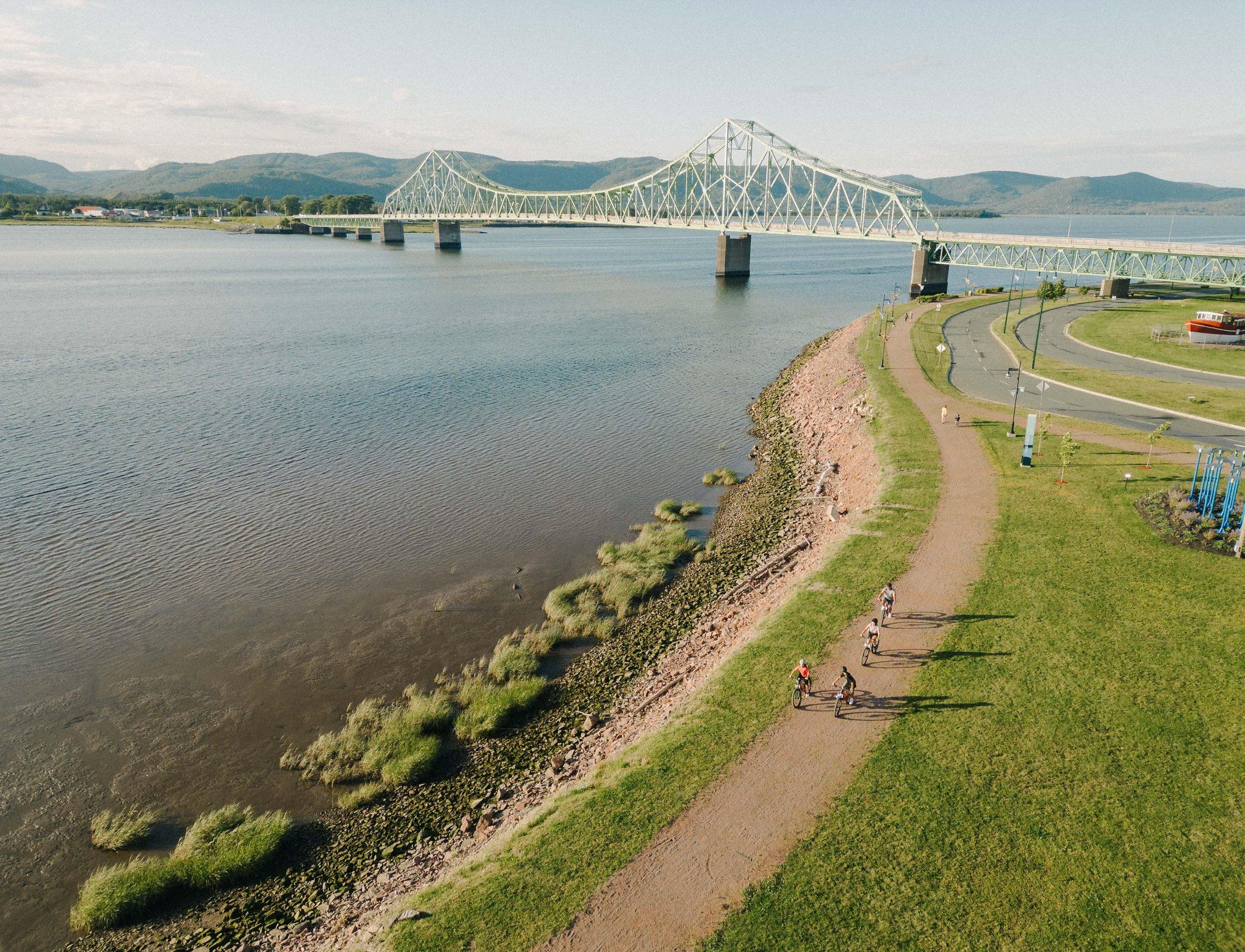 Een pad langs de rivieroevers, langs kalm water met in de verte een hangbrug en groene heuvels onder een heldere hemel.
