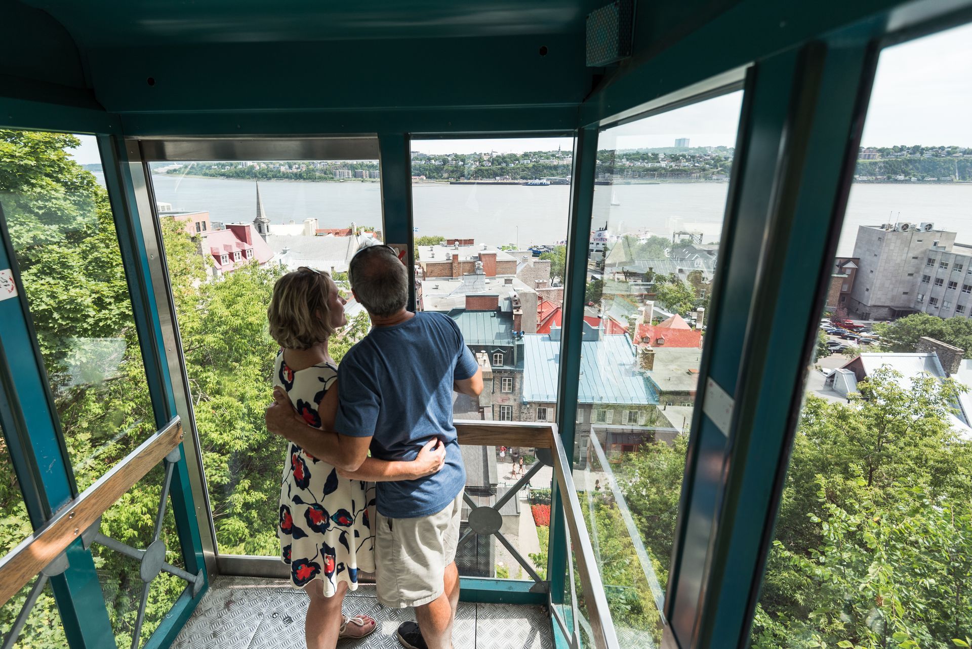 Een persoon die een kind vasthoudt in een glazen kabeltrein, met uitzicht over Old Quebec en de haven.