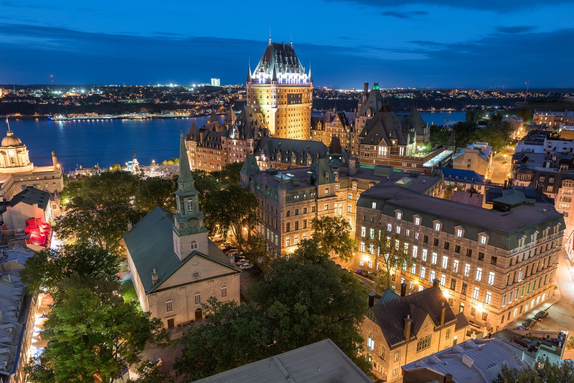 Nachtelijk stadsgezicht van Old Quebec met een fel verlicht historisch gebouw aan een rivier en havenlichten in de verte.