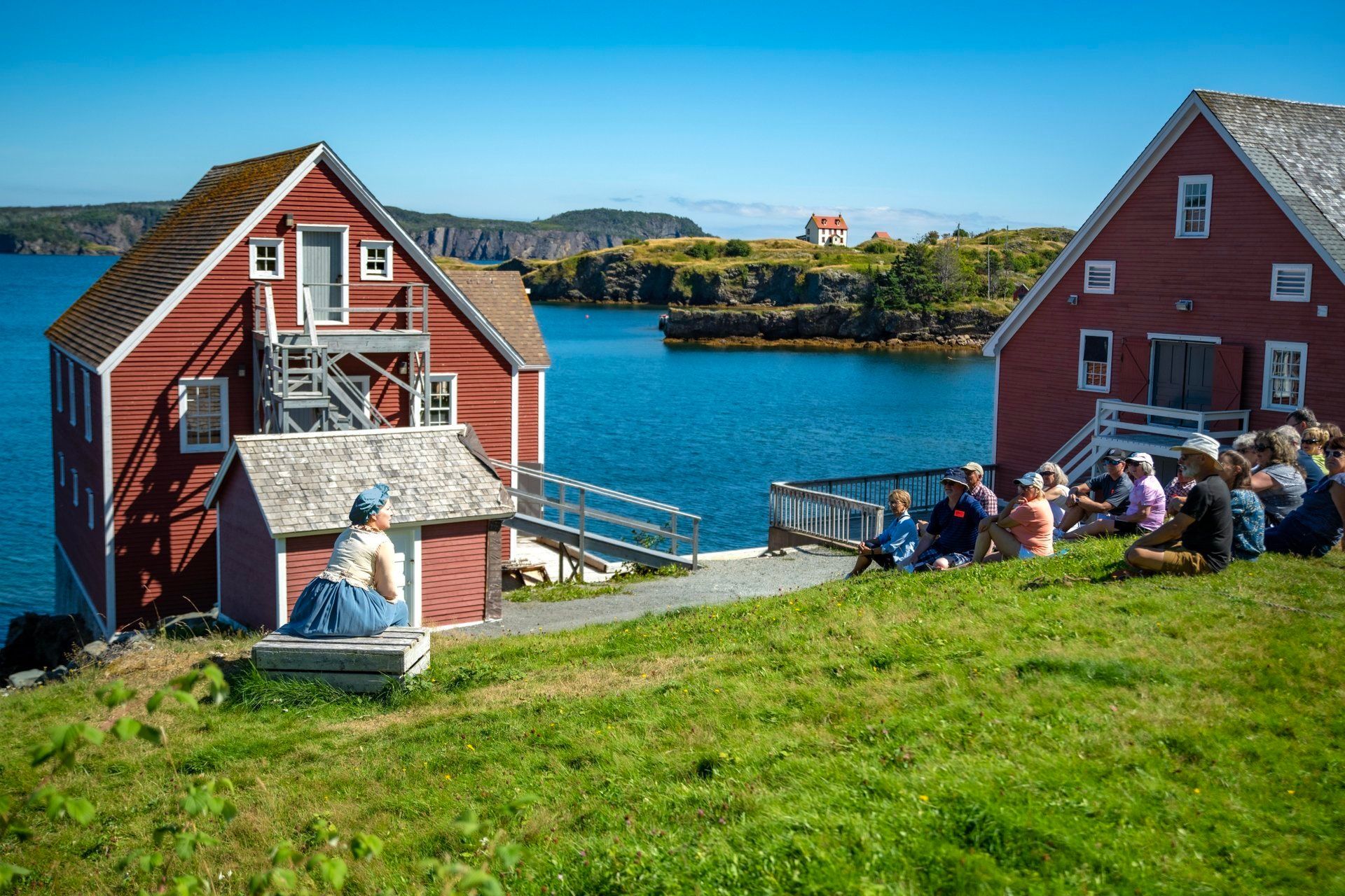 Rode huizen van Trinity in Newfoundland, Canada, aan de waterkant en een zittende menigte die uitkijkt over een blauwe haven op een zonnige dag.