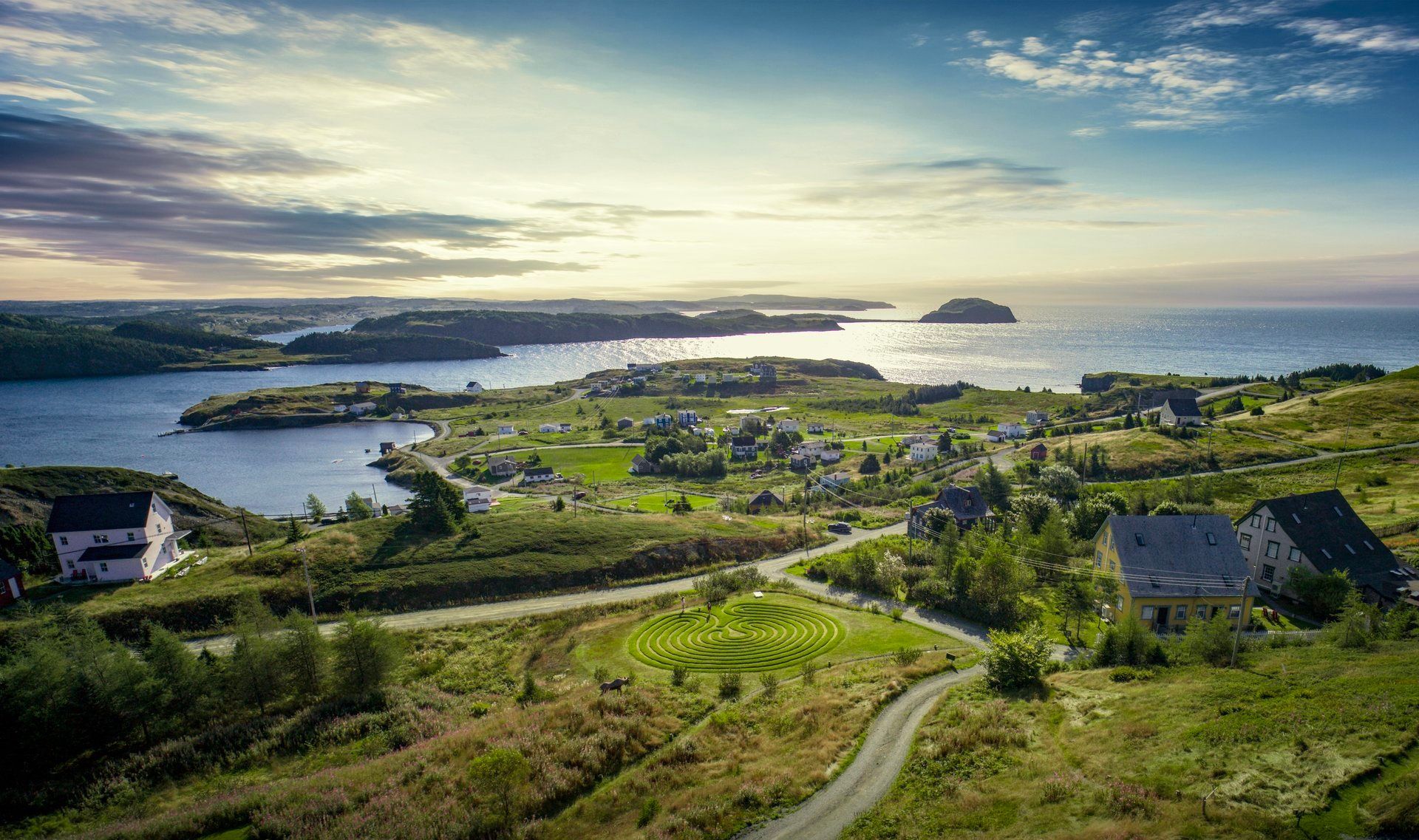 Het kustdorp Port Rexton, Newfoundland Canada, met kronkelende wegen, groene heuvels en een kalme zee bij zonsondergang onder een bewolkte hemel.