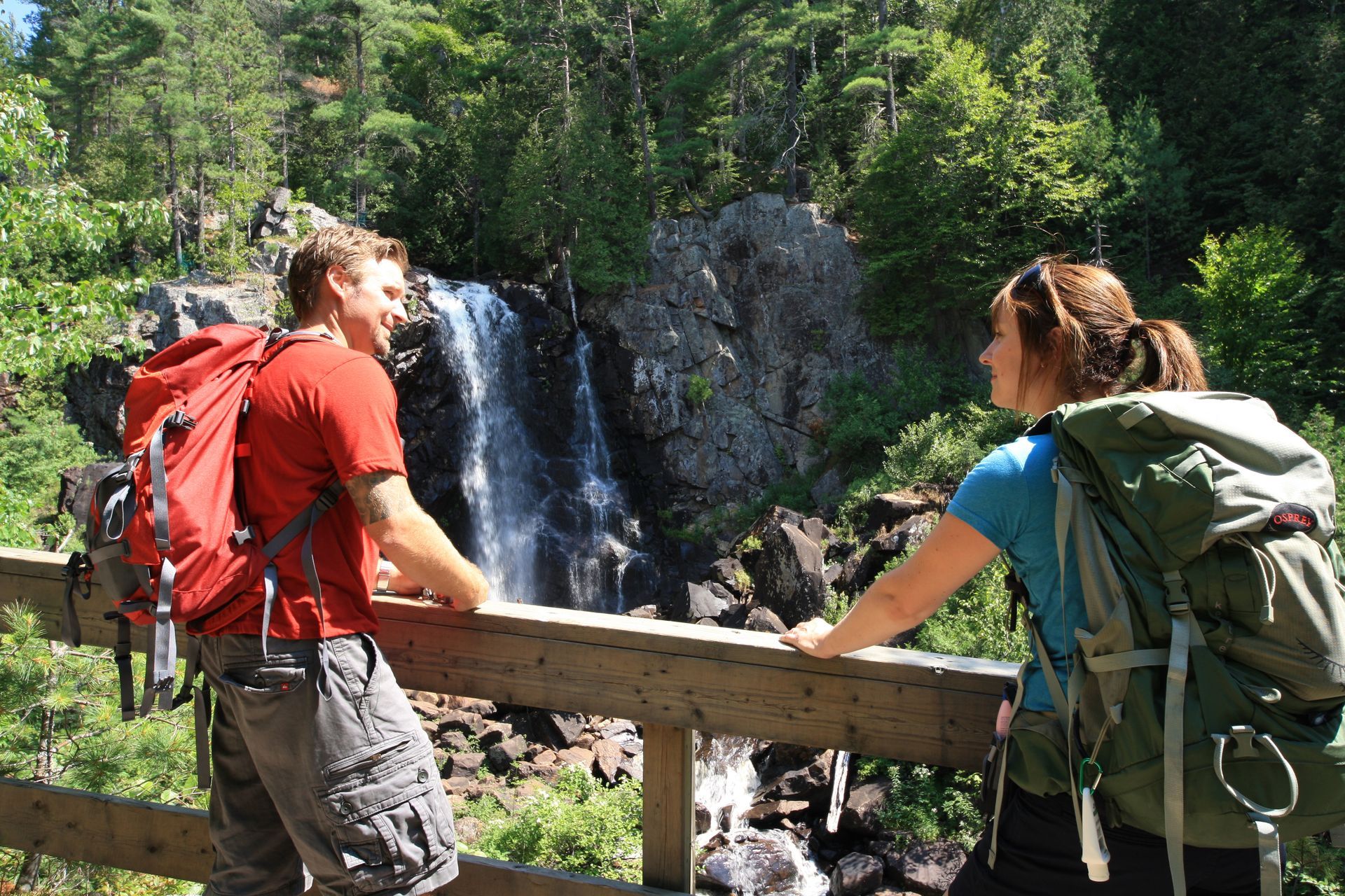 Twee wandelaars in Canada met rugzakken pauzeren op een houten brug naast een waterval in een bos.