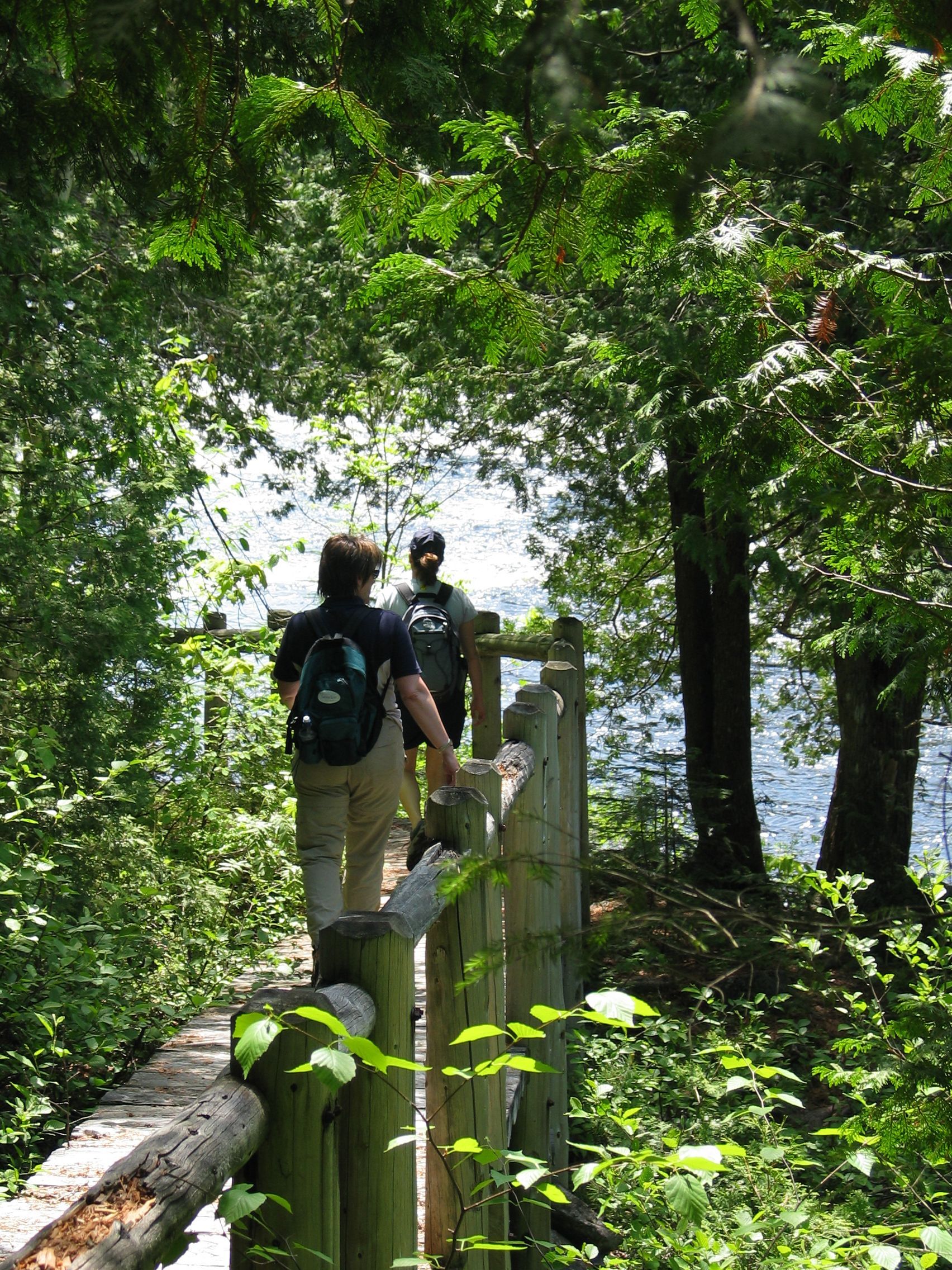 Twee wandelaars in Canada lopen over een smal bospad langs een houten hek en het water.