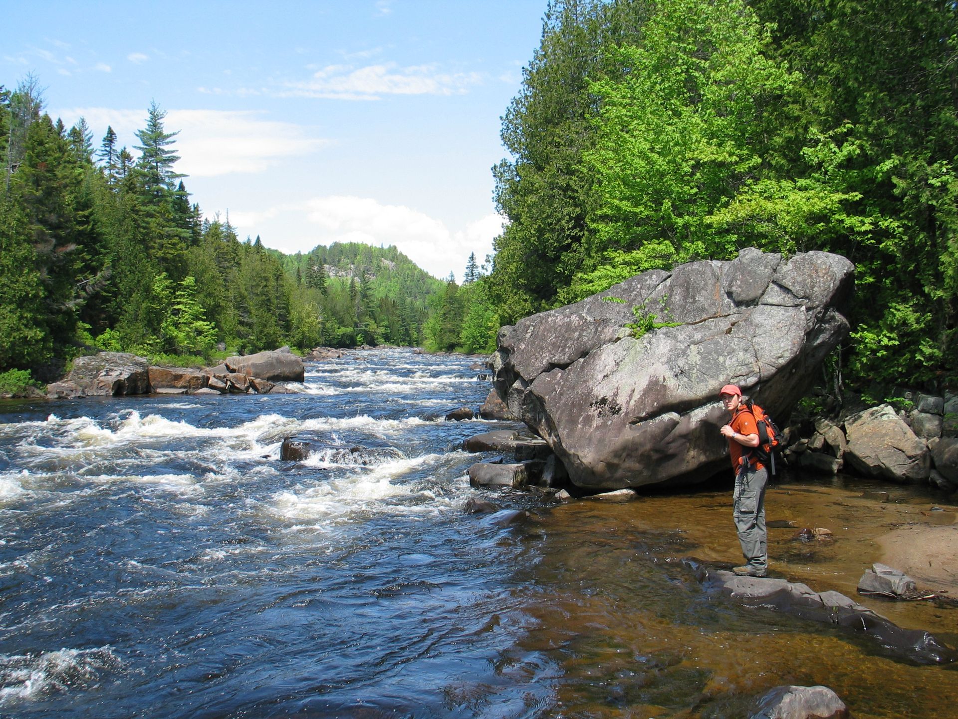 Persoon in oranje staat bij een rotsachtige rivier in Canada in een bosrijk landschap.