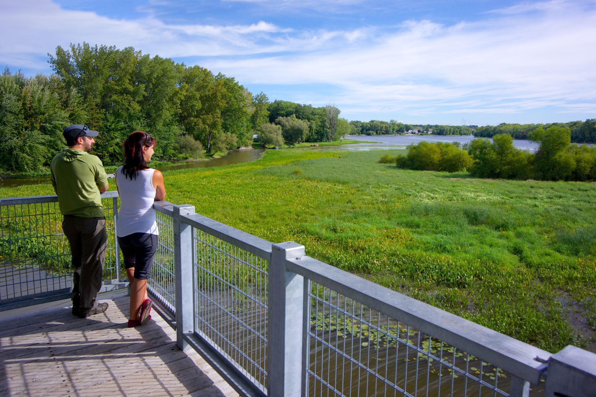 Twee mensen staan op een houten vlonderpad in Canada met uitzicht op een groen moerasgebied en een rivier onder een bewolkte hemel.