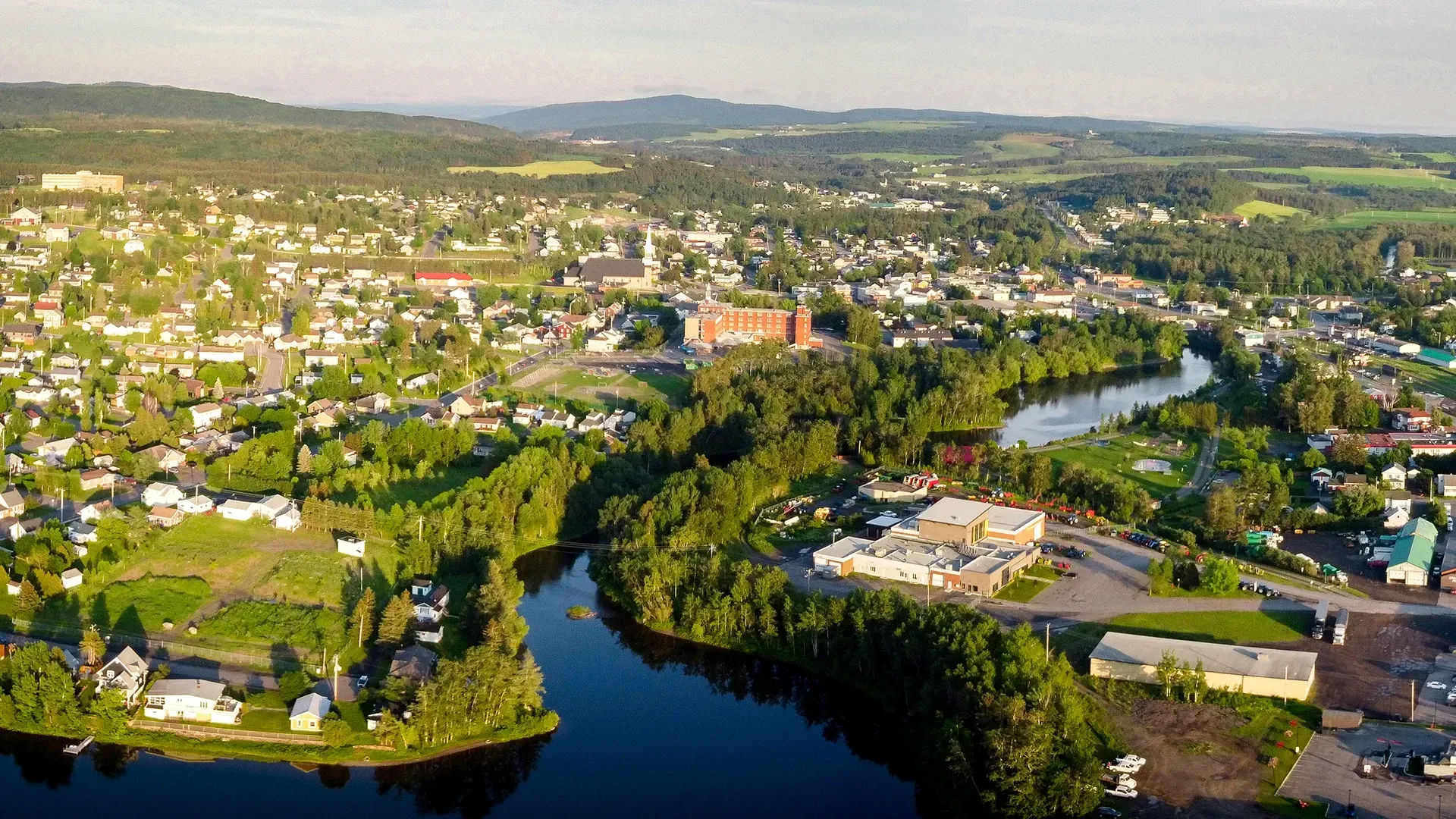 Luchtfoto van het stadje Amqui in Quebec, Canada, aan een kronkelende rivier, met groene heuvels en verspreide gebouwen in de verte.