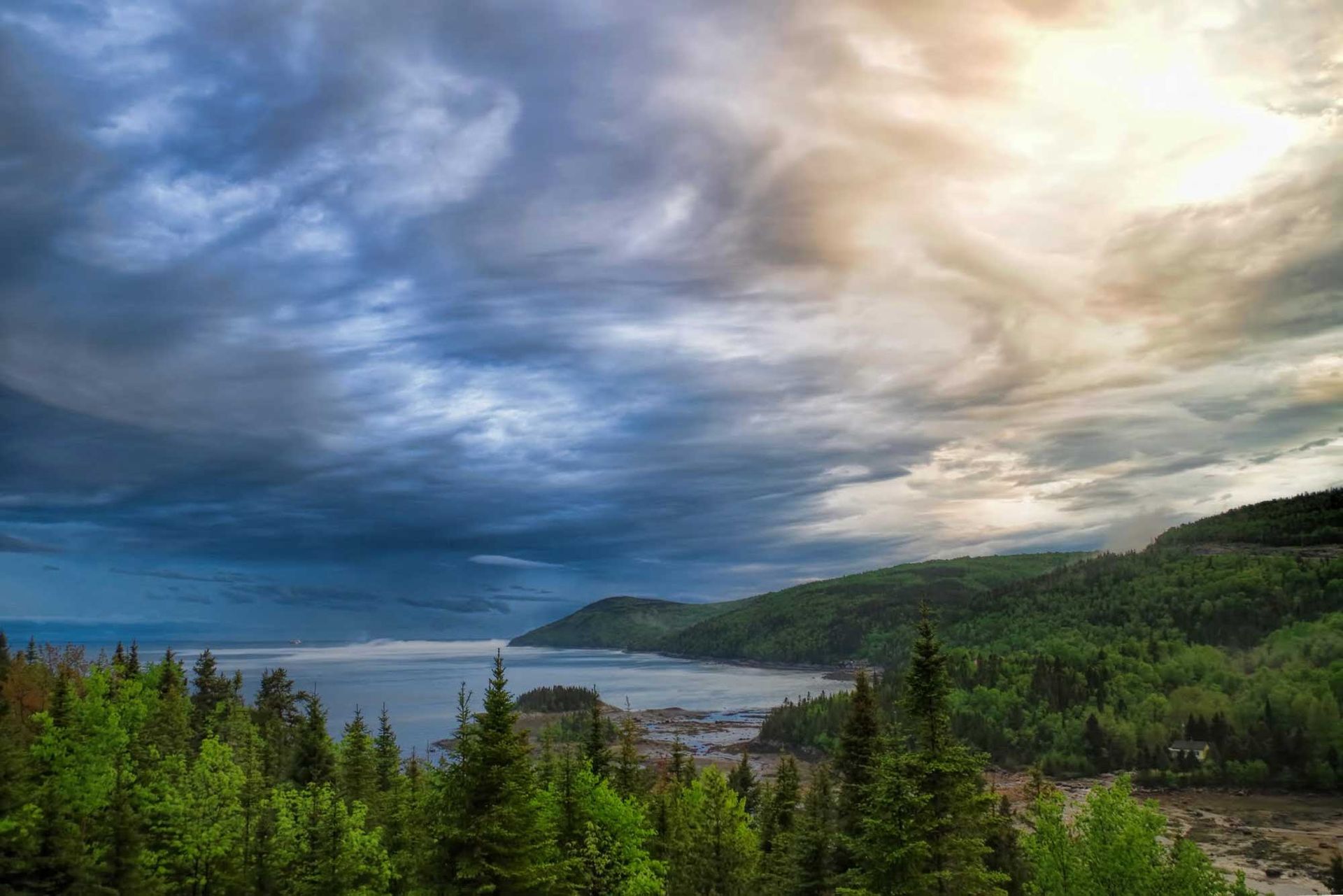 Een bewolkt kustlandschap in Quebec, Canada, met beboste heuvels, blauw water en de zon die door een dramatische hemel breekt.