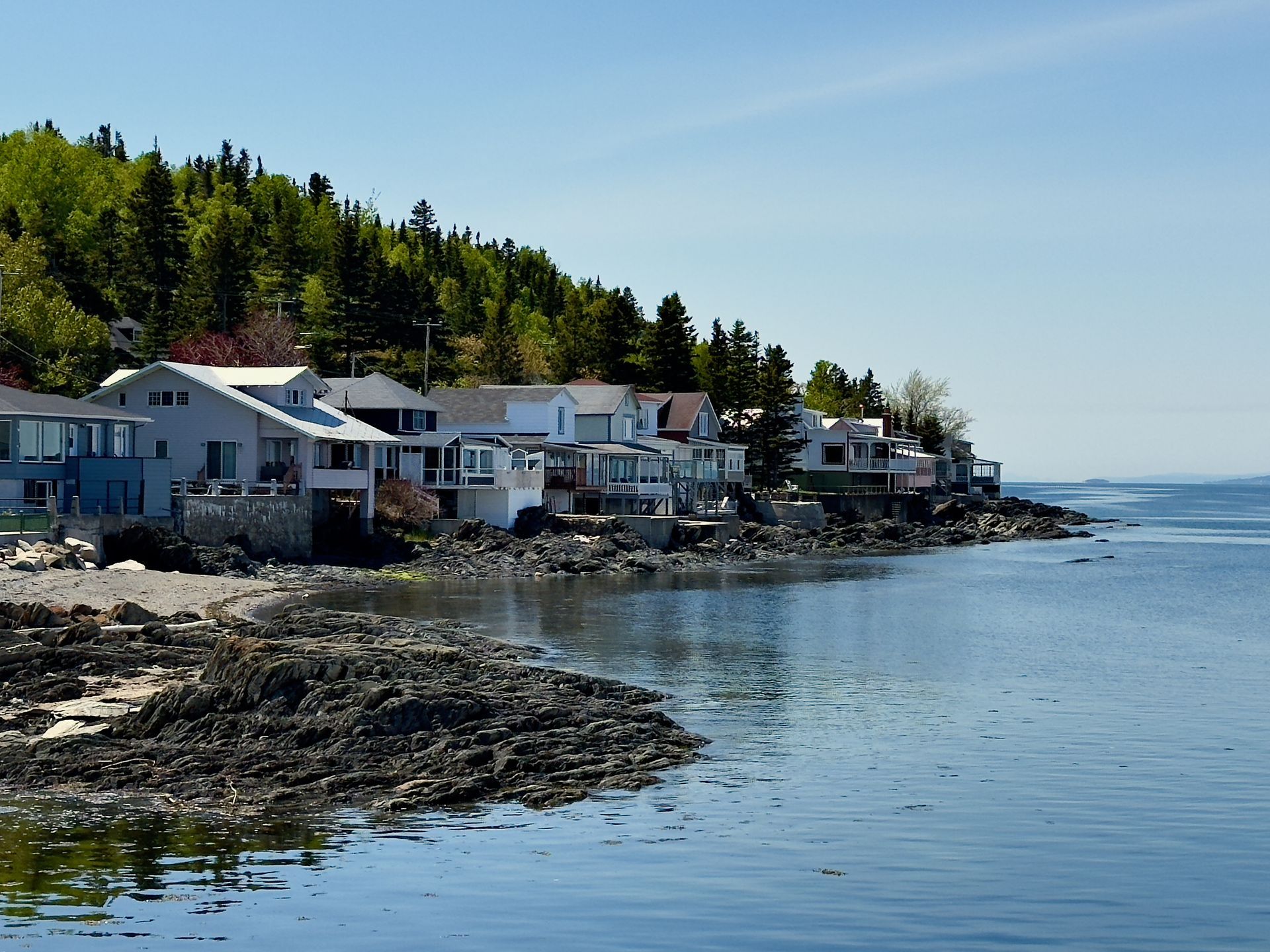 Een rotsachtige kustlijn in Trois-Pistoles, Quebec Canada, met witte huizen, groene bomen en kalm blauw water onder een heldere hemel.