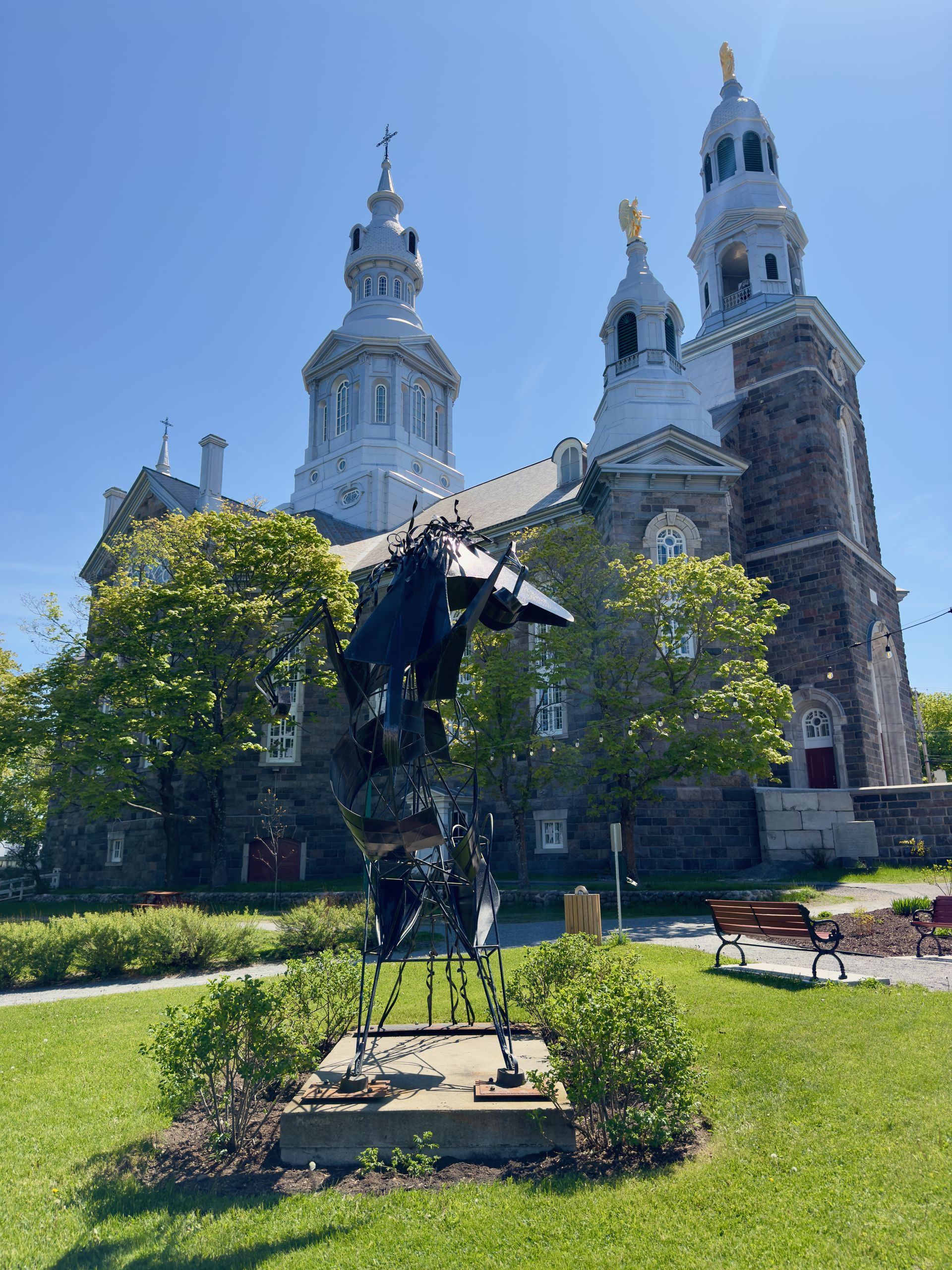 Bronzen beeld in Trois-Pistoles, Quebec Canada,,in een zonnig park voor een kerk met een witte koepel, twee torens en een groen gazon.