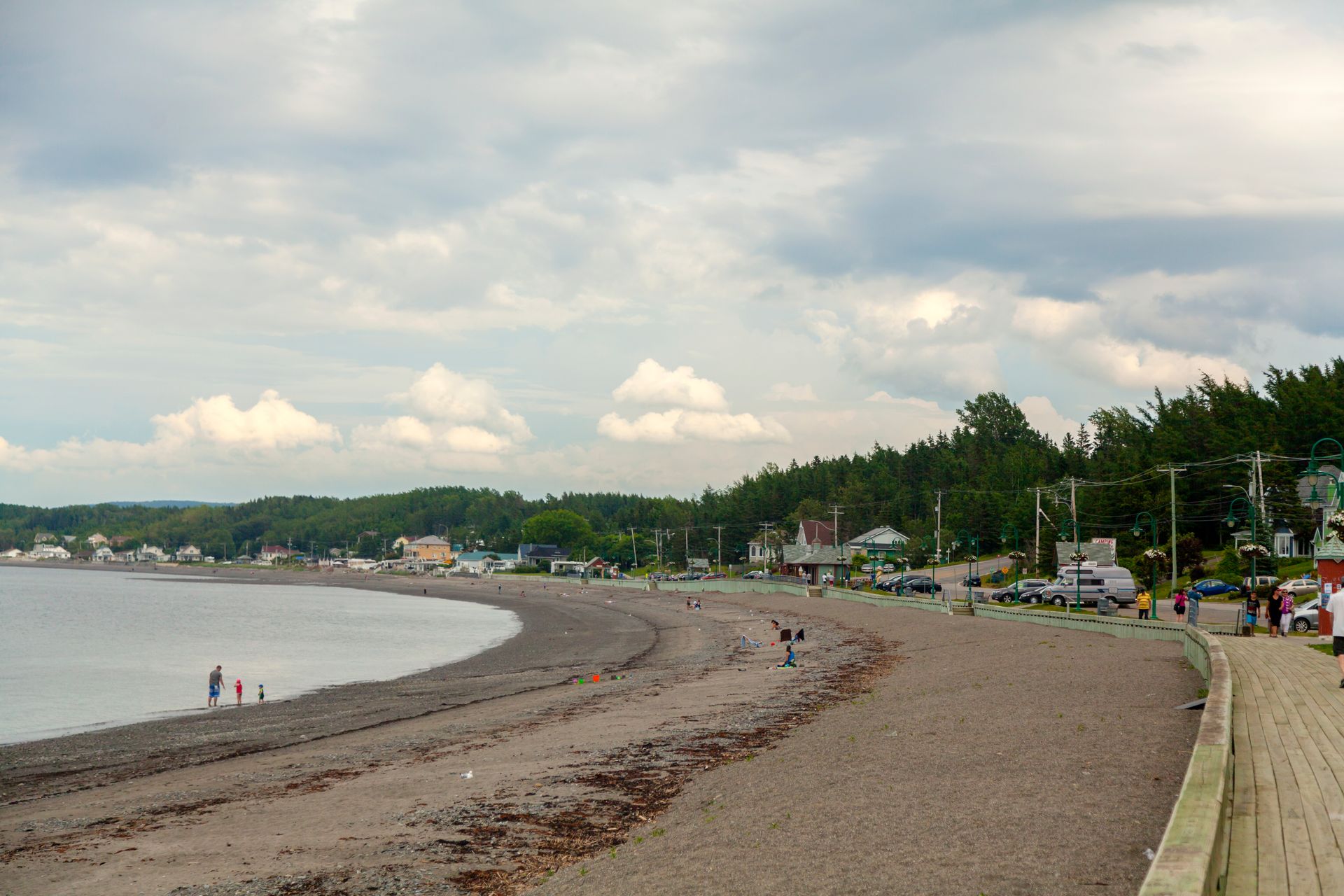 Een breed zandstrand an de Saint Lawrence River bij Sainte-Luce-sur-Mer, Quebec Canada, met weinig mensen, kalm water en een met bomen omzoomde kustlijn onder een bewolkte hemel.