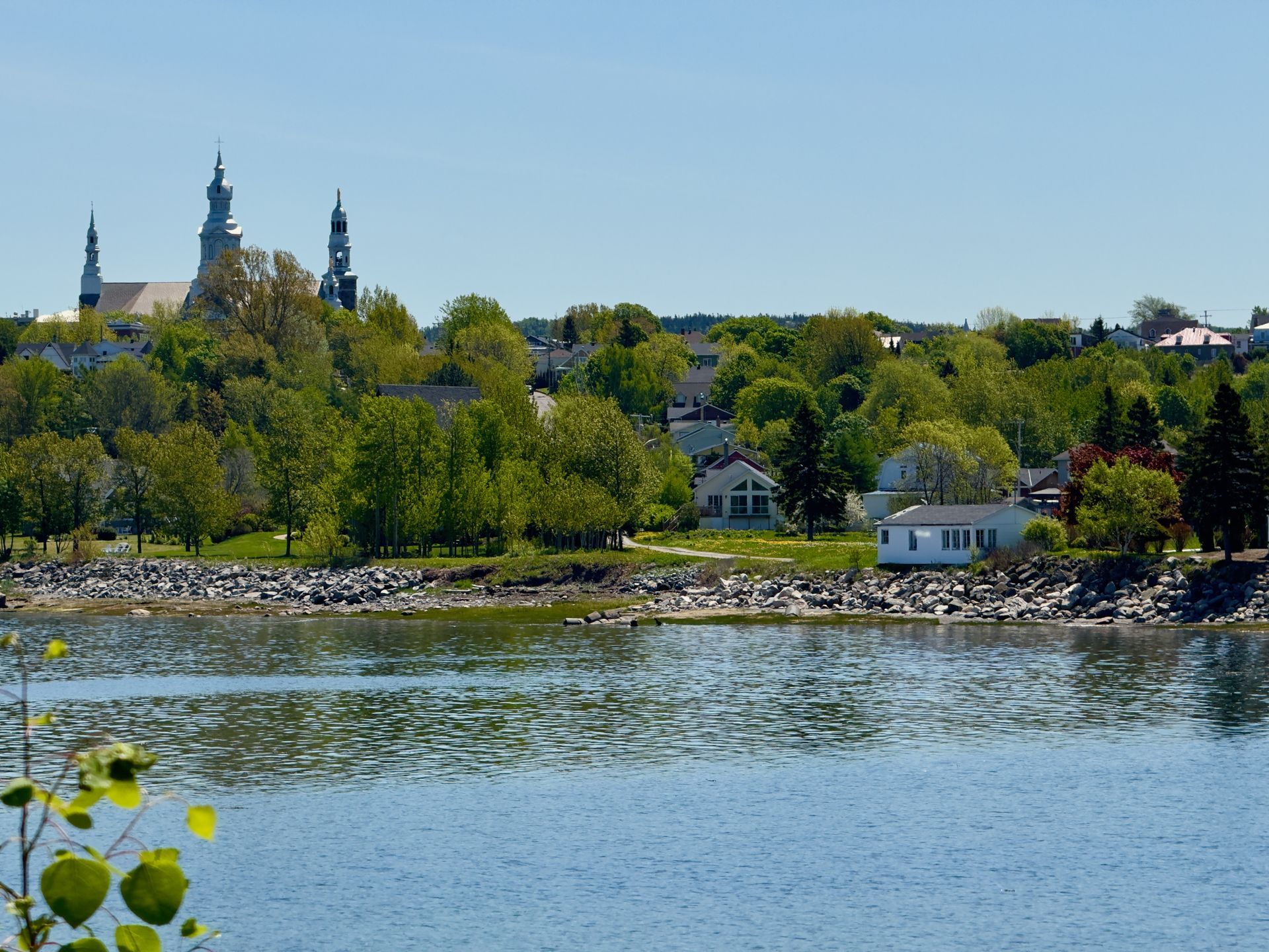 De skyline van Trois-Pistoles in Quebec, Canada, aan het water met kerktorens boven de bomen en kalm blauw water op de voorgrond.