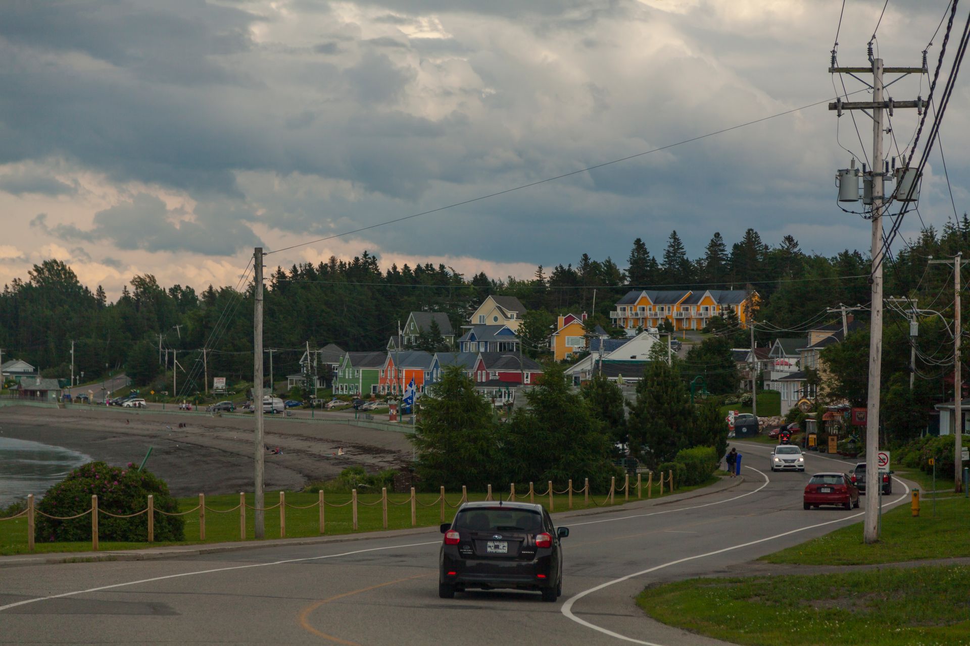 Een bochtige weg bij De weg bij Sainte-Luce-sur-Mer , Qubec Canada, met meerdere auto's langs een met bomen omzoomde heuvel in een woonwijk, onder een bewolkte hemel.