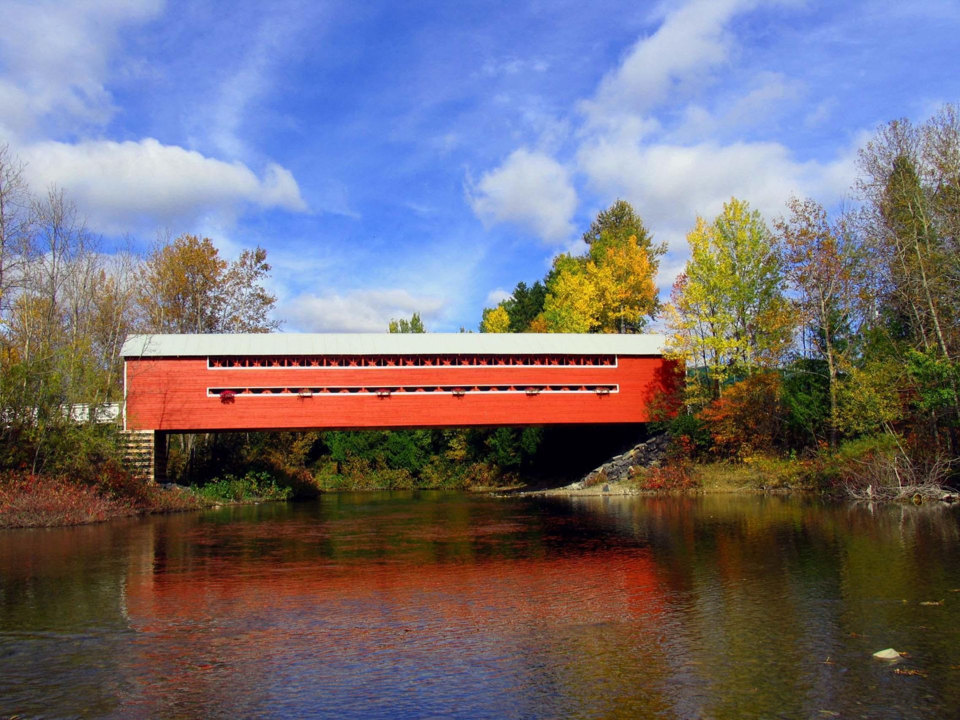 Een rode Covered Bridge in Amqui, Quebec Canada, over een rustige rivier, omlijst door herfstbomen en een blauwe hemel.