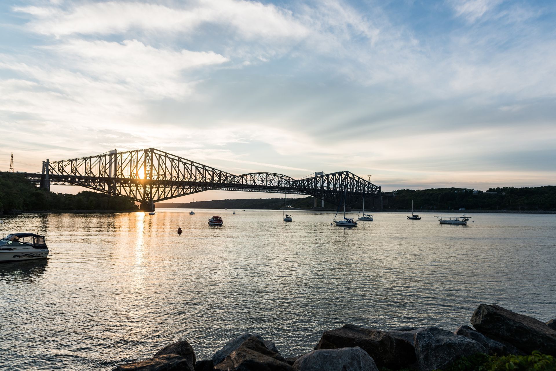 Zonsondergang boven de Quebec Bridge die over kalm water loopt, met kleine bootjes en een rotsachtige kustlijn op de voorgrond.