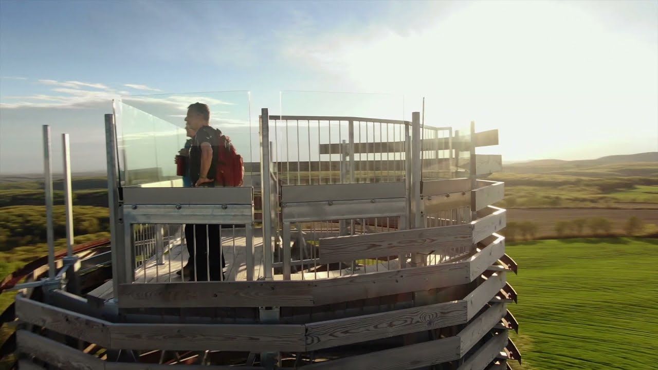 Een persoon staat op een rond houten uitkijkplatform bij zonsondergang, met een open landschap op de achtergrond.