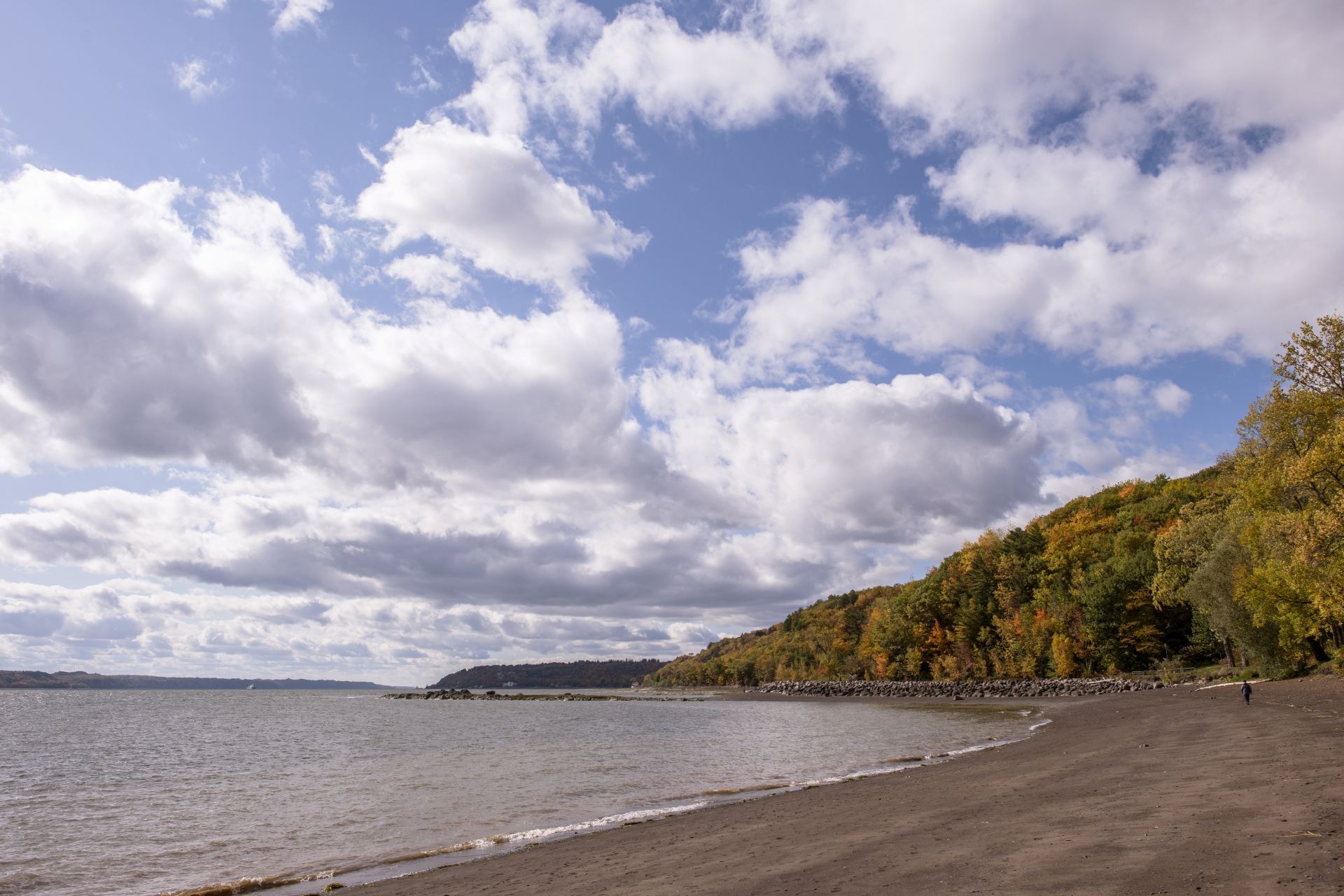 Een kiezelstrand in Quebec, Canada, aan kalm water onder een bewolkte hemel, met aan de rechterkant een met bomen begroeide heuvel.