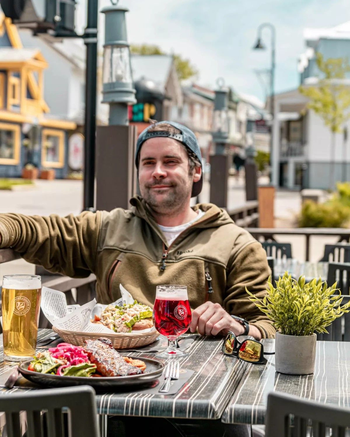 Man aan een tafeltje op een terras in Baie-Saint-Paul in Charlevoix, Canada, met bier, salade en een rode drank in een zonnige straatomgeving.