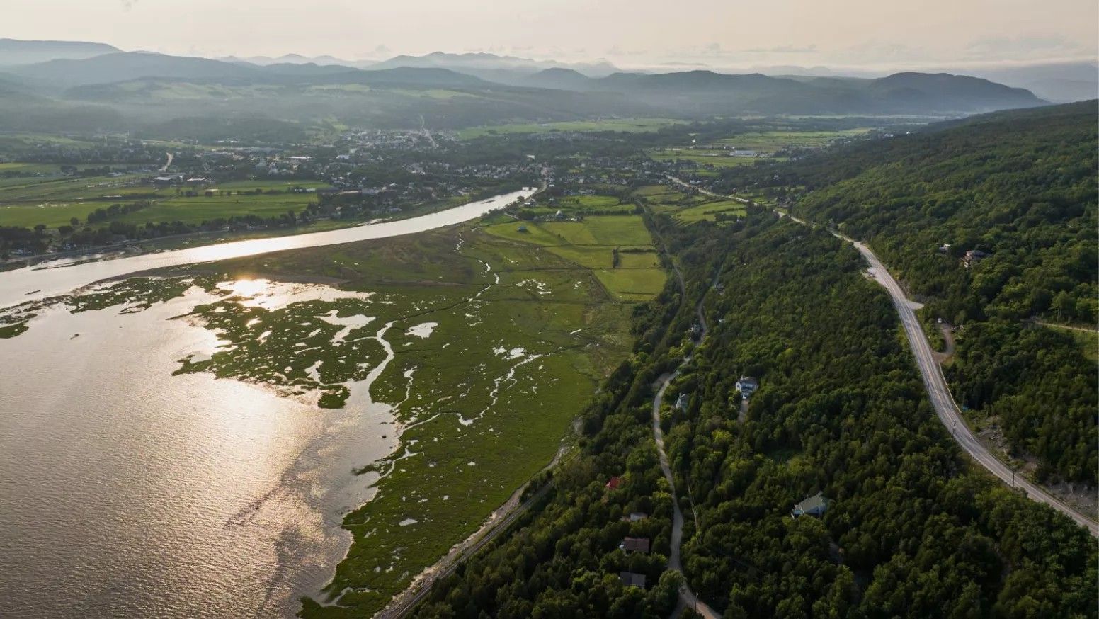 Luchtfoto van Baie Baie-Saint-Paul in Charlevoix, Canada, met groene heuvels, een kronkelende weg en zonovergoten water dat de lucht weerspiegelt.