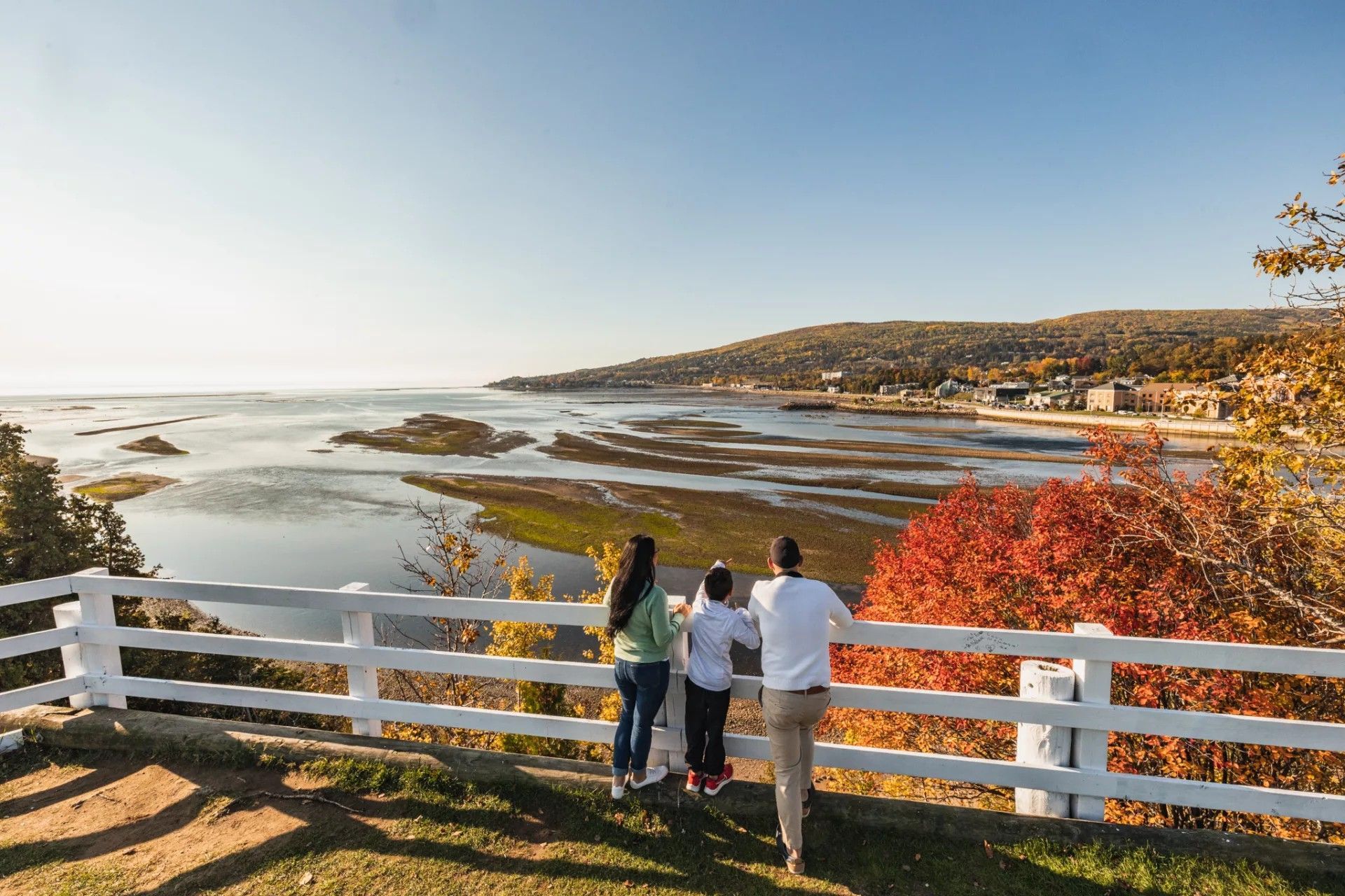 Drie mensen kijken vanaf een wit hek uit over een rotsachtige kustlijn en een kalme zee bij zonsondergang bij Cap-à-l'Aigle in Quebec, Canada.