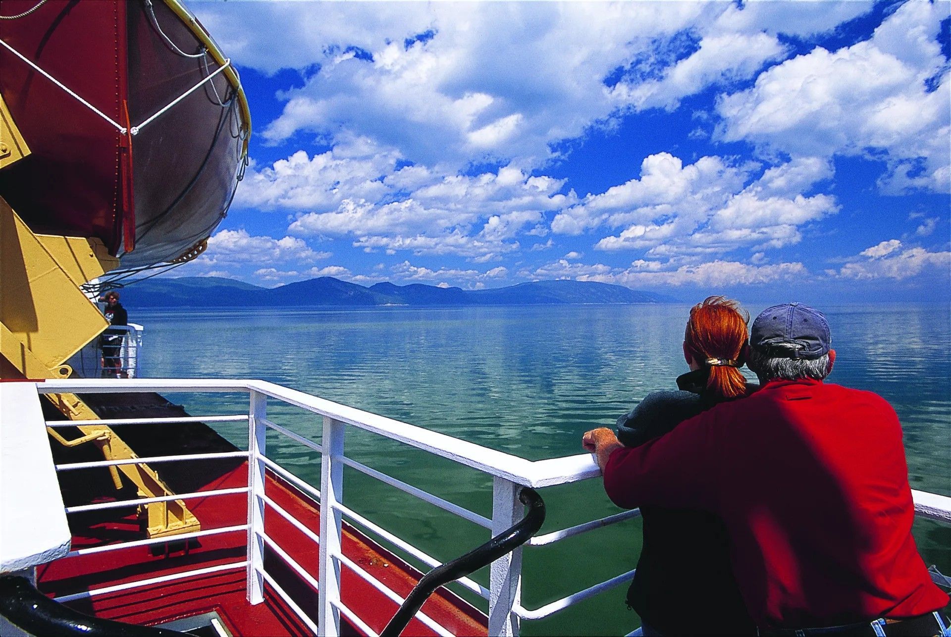 Twee mensen in rode jassen staan op het dek van een boot en kijken uit over de St.Lawrence Rivier in Quebec, Canada, en de bergen in de verte.