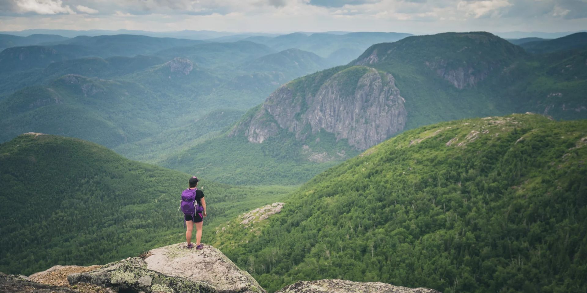 Een wandelaar in een paarse outfit staat op een rotsachtige klif van Parc national des Grands-Jardins in Canada met uitzicht op een weelderig groene bergvallei en verre kliffen.