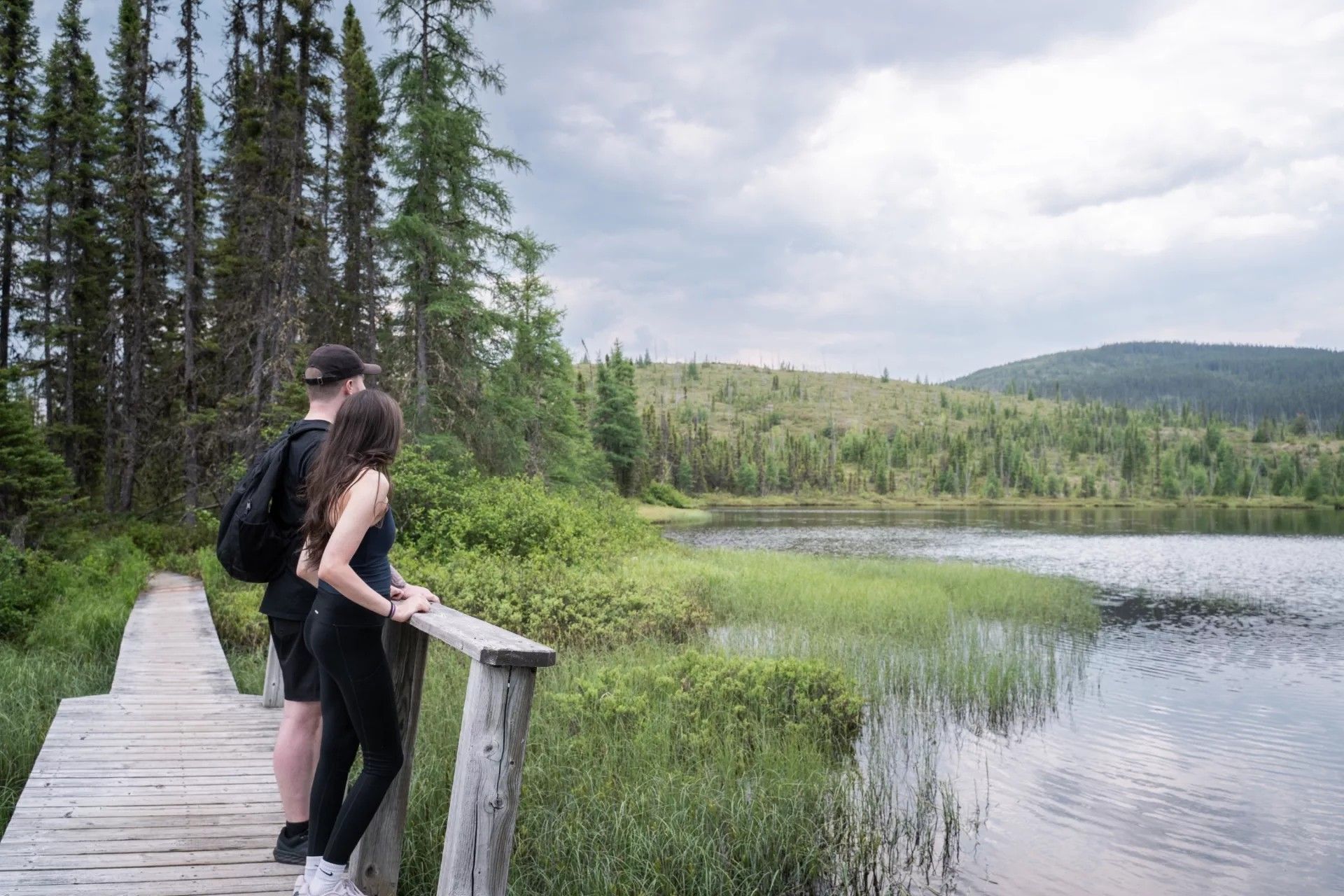 Twee mensen op een houten vlonderpad van Parc national des Grands-Jardins in Canada met uitzicht op een rustig meer en een dennenbos onder een bewolkte hemel.
