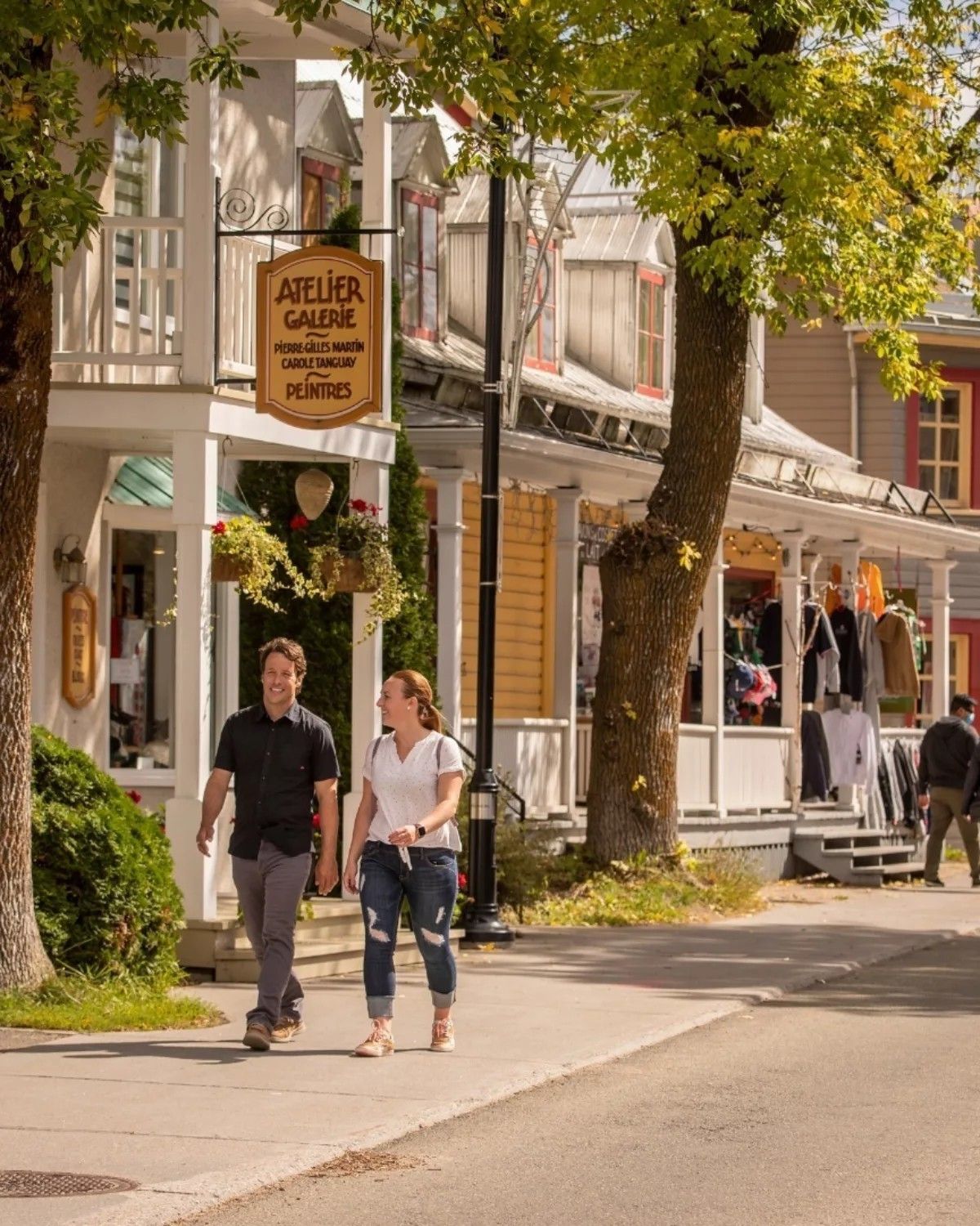 Mensen wandelen over een zonnige straat in Baie-Saint-Paul in Charlevoix, Canada, langs historische winkels en een cafébord.