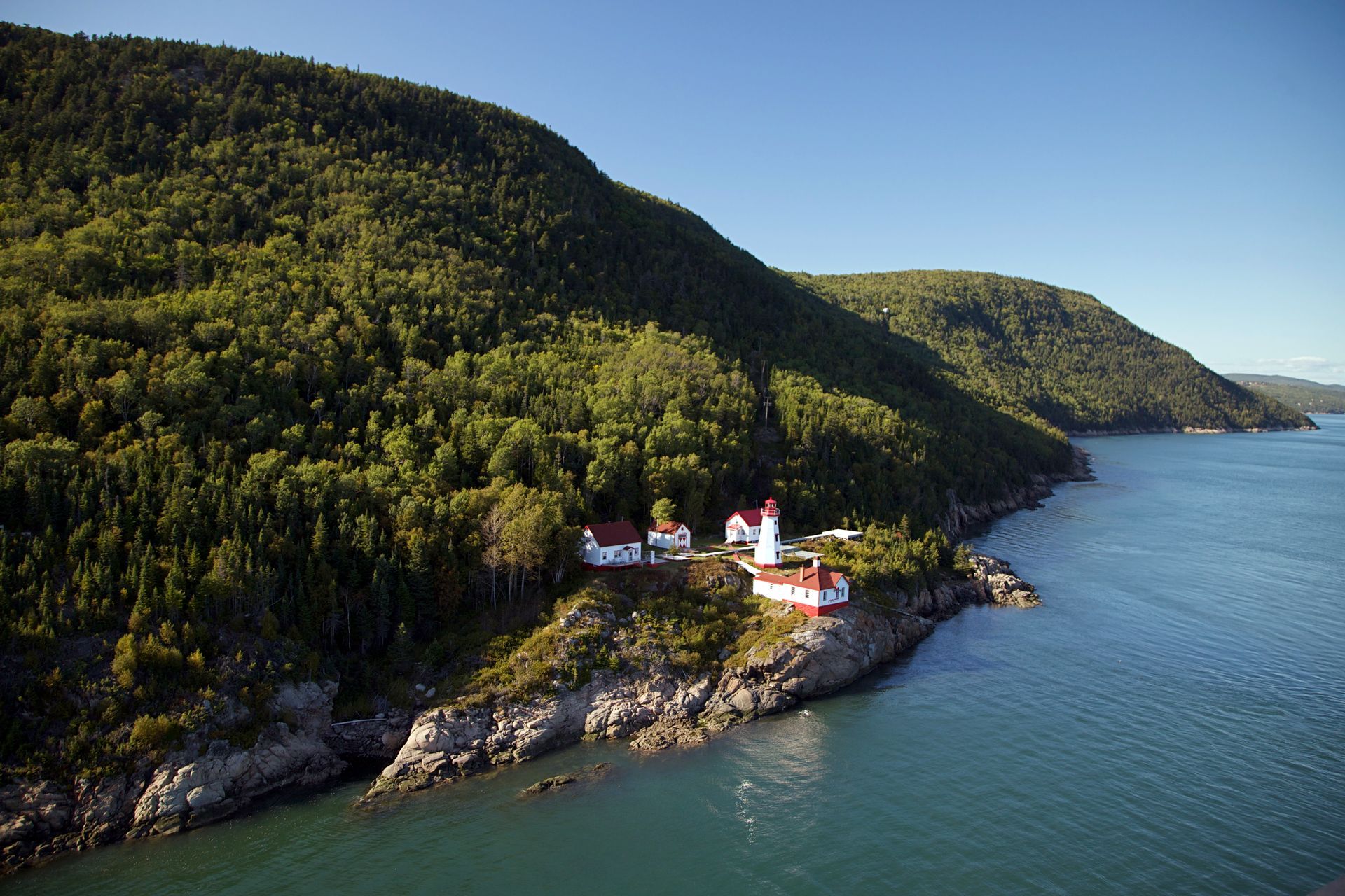 De rood-witte McLaren Chapel in Port-au-Persil in Quebec, Canada, op een rotsachtige, beboste kustlijn aan kalm blauw water.