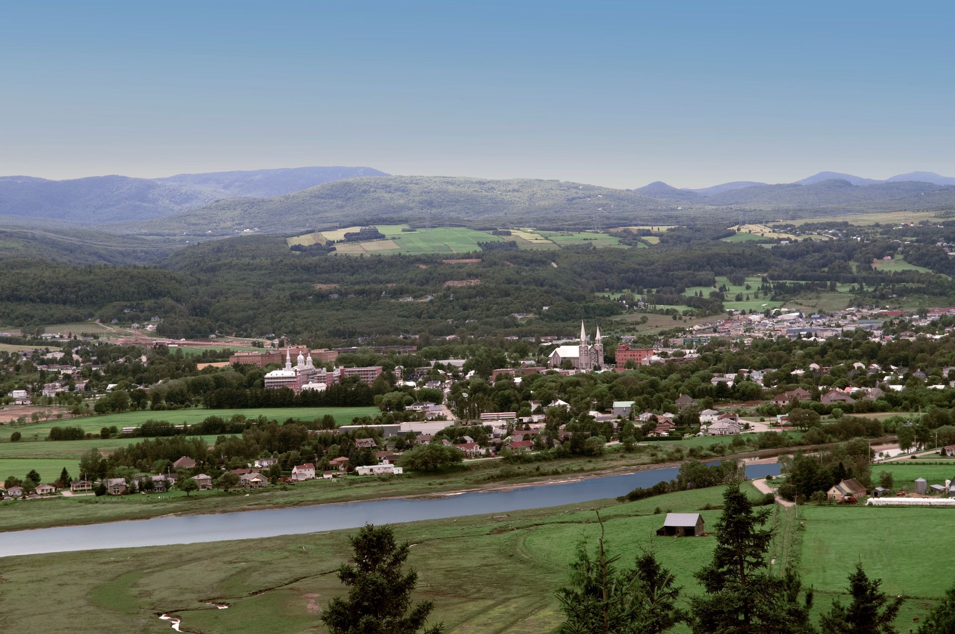 Het uitgestrekt dalstadje Baie-Saint-Paul in Canada met een rivier, groene velden en in de verte bergen onder een nevelige hemel.