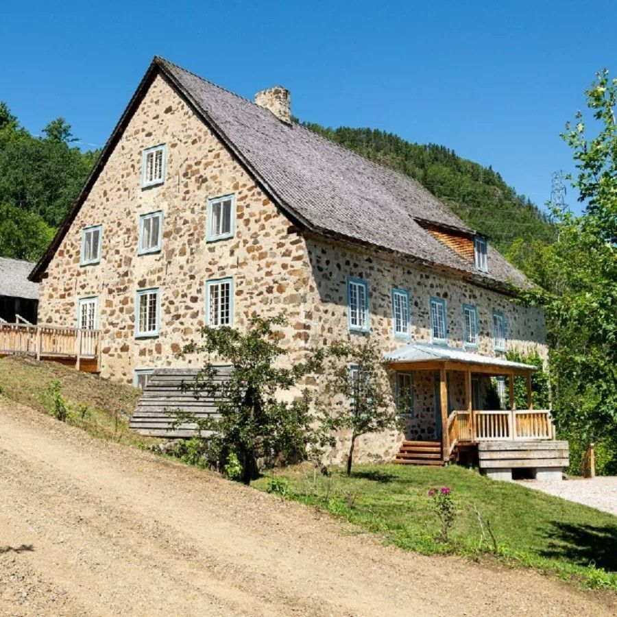 Moulin de la Rémy in Charlevoix, Canada, met een steil rieten dak op een zonnige heuvel, omgeven door bomen en groen.