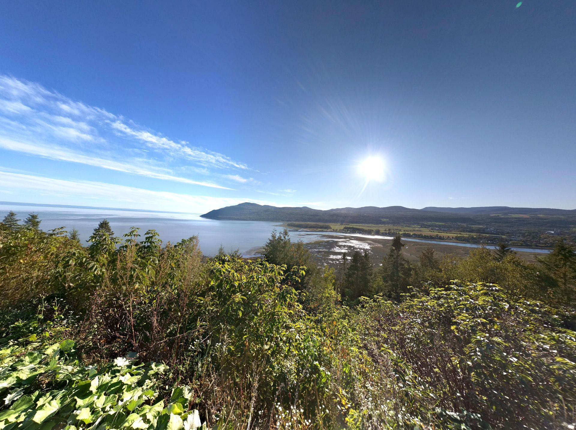 Zonnig uitzichtpunt aan de kust nabij Baie-Saint-Paul in Canada met blauwe lucht, oceaan, heuvels en groene struiken op de voorgrond.