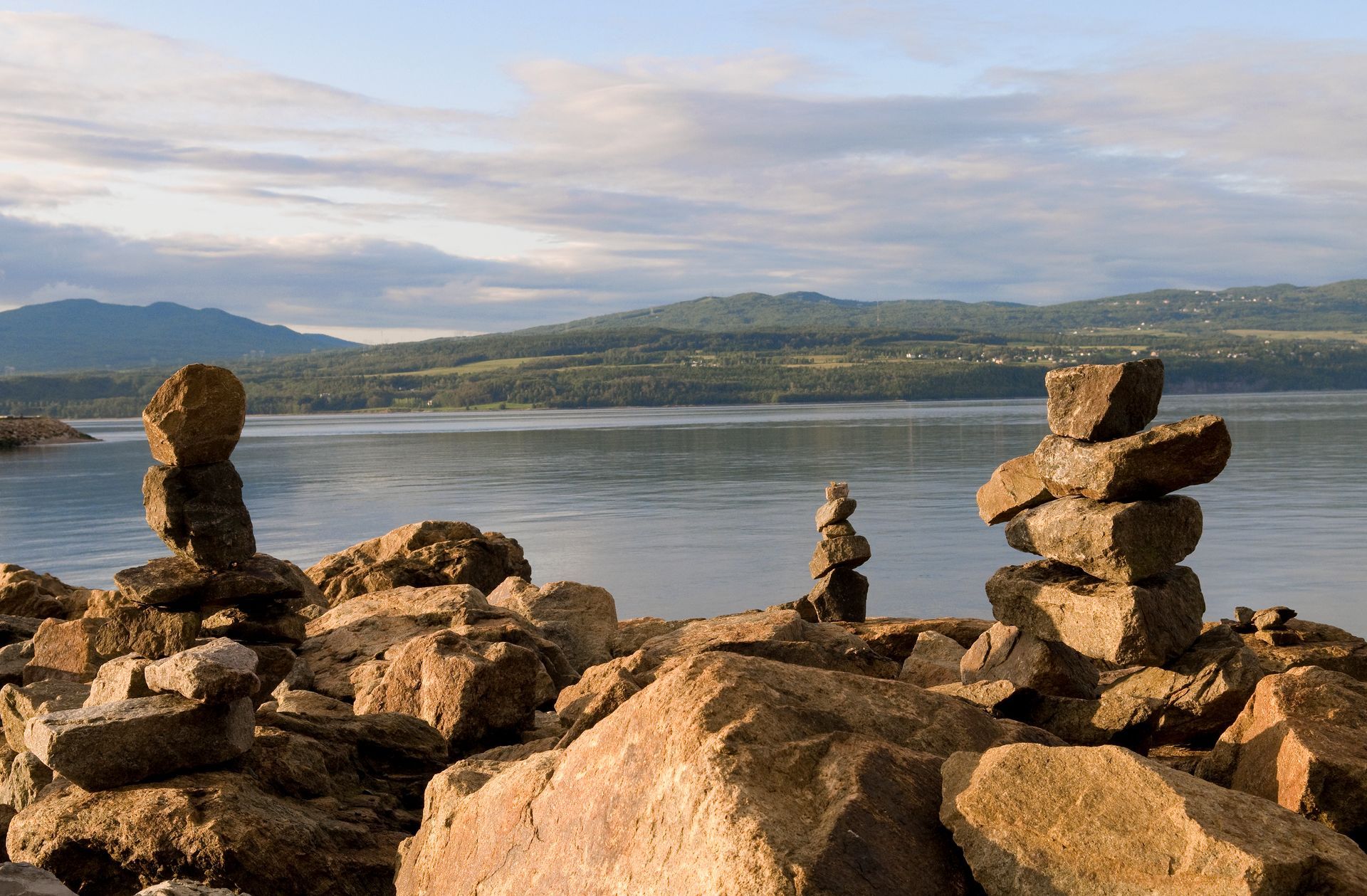 Steenstapels in Quebec, Canada, op een rotsachtige oever naast een kalm meer met bergen onder een bewolkte hemel.