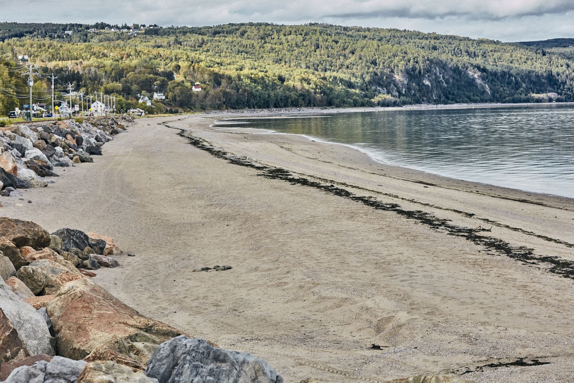 Het zandstrand van Saint-Irénée in Quebec, Canada, met rotsen, kalm water en een met bomen begroeide kustlijn onder een heldere hemel.