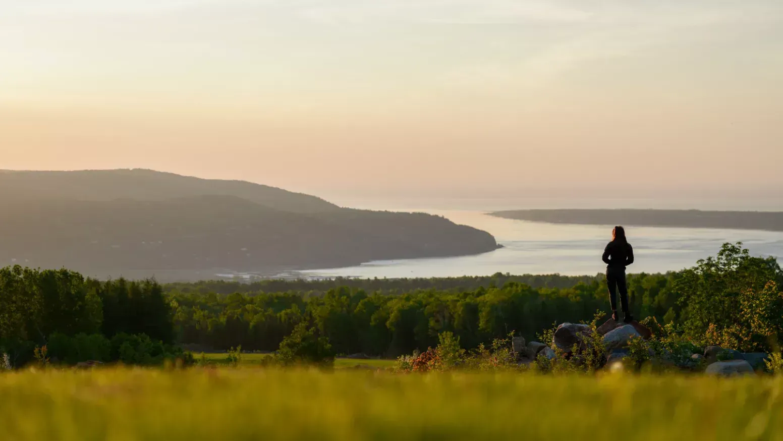 Een persoon staat op Belvédère de Baie-Saint-Paul in Charlevoix, Canada, met uitzicht op een kalme baai bij zonsondergang.