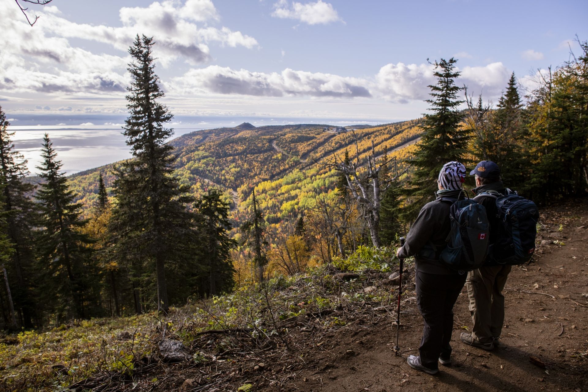Twee wandelaars met rugzakken op een bospad nabij Saint-Tite-des-Caps in Canada met uitzicht op een zonovergoten bergdal.