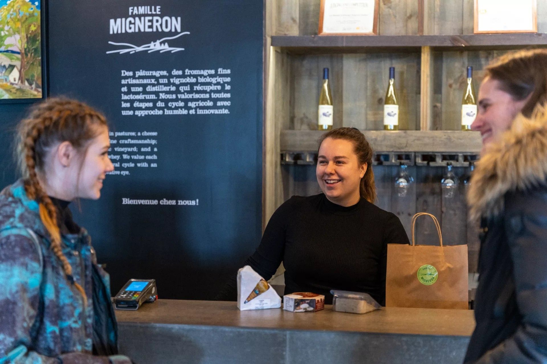 De balie van Famille Migneron de Charlevoix in Canada met twee lachende medewerkers en een klant, schappen en een krijtbordmenu erachter.