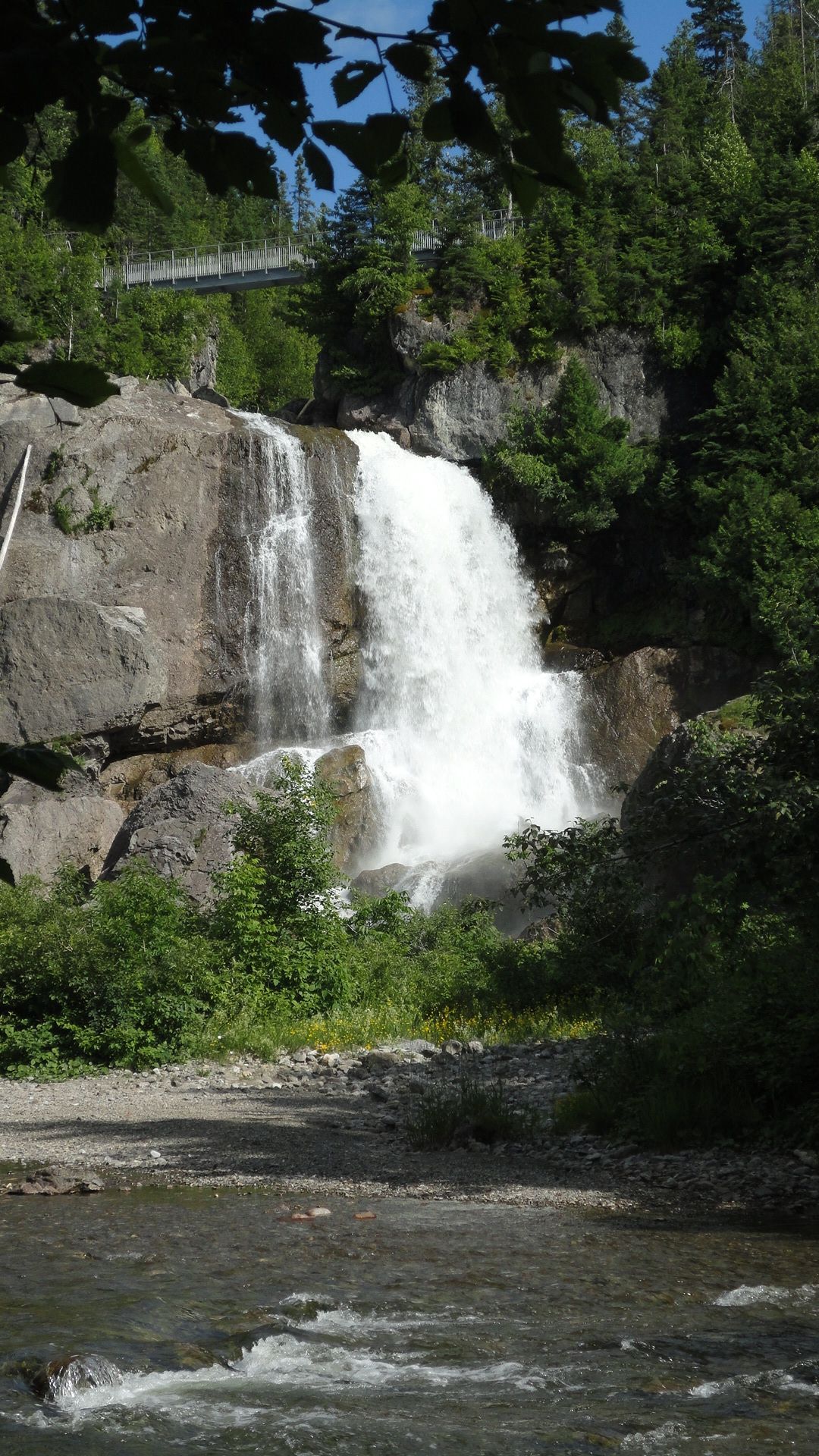 Een waterval in Quebec, Canada, die over een rotsachtige klif naast een rivier naar beneden stort, met groene bomen en een brug erboven.