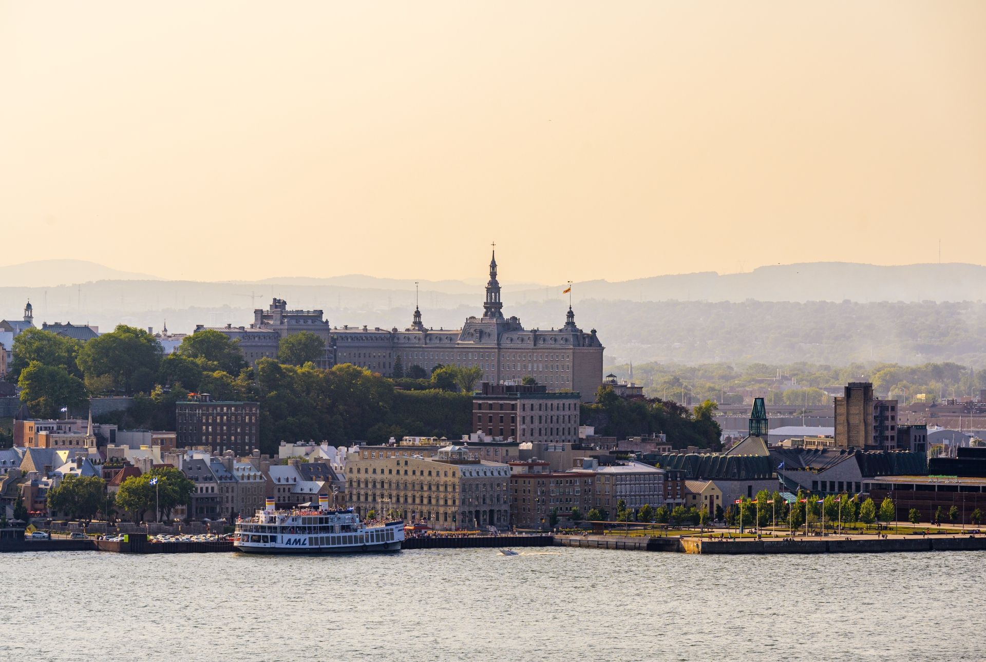 De skyline van Quebec bij zonsondergang, met boten op het kalme water en een kasteel op een heuvel in de verte.